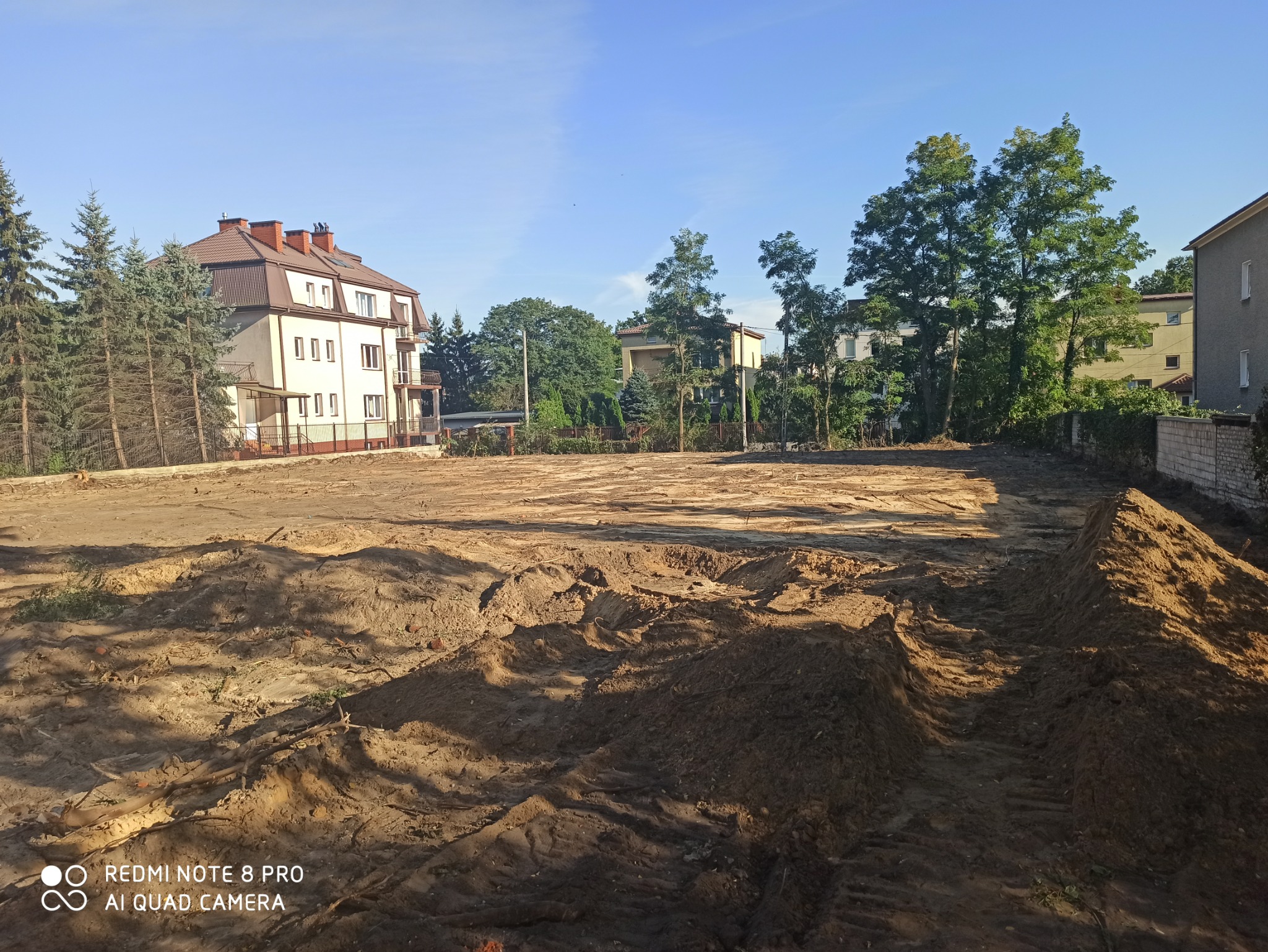 Przygotowany teren pod budowę z widocznymi śladami opon maszyn budowlanych, w tle budynki mieszkalne i drzewa.