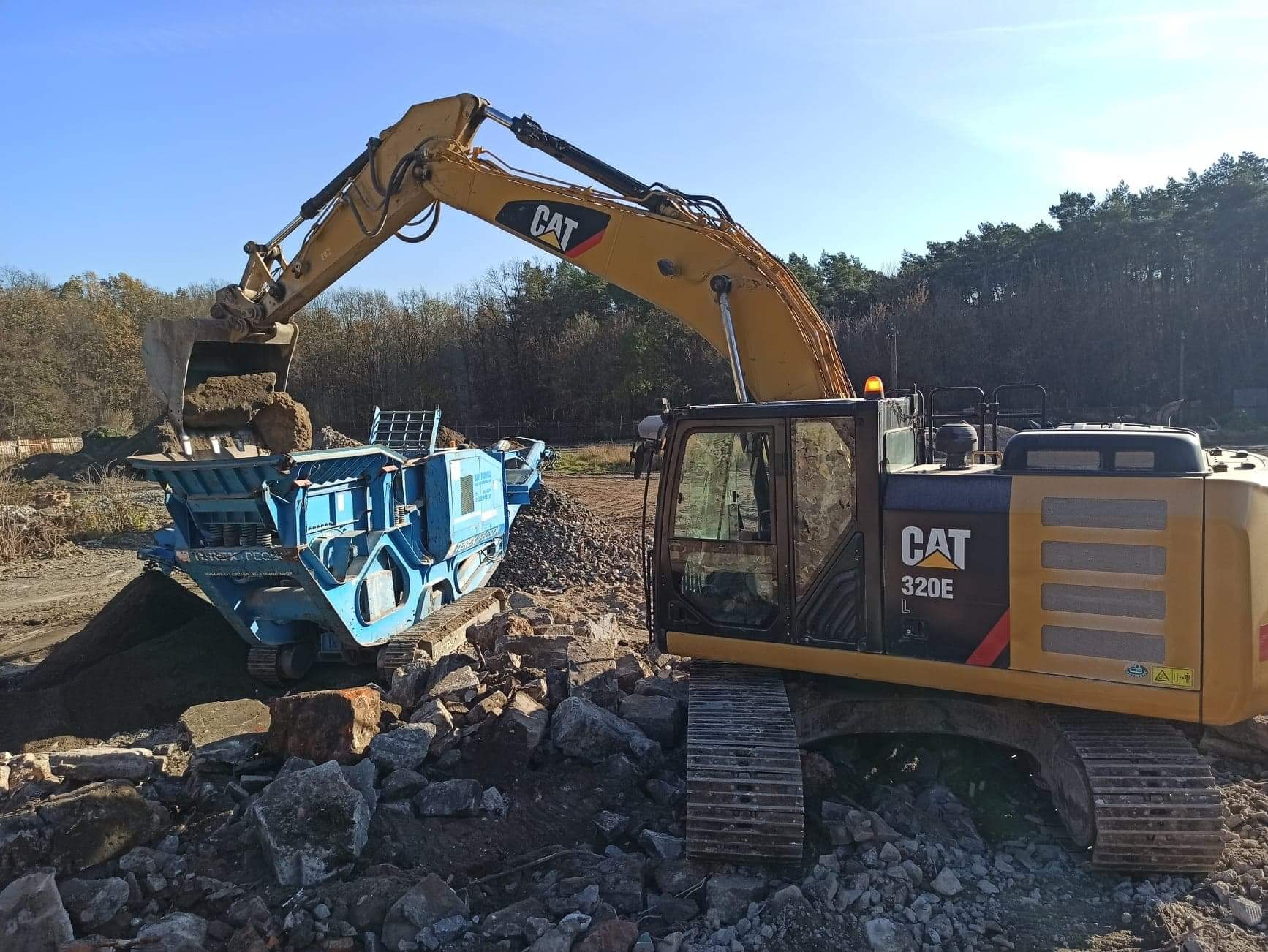 Żółta koparka CAT 320E z łyżką załadowuje urobek do niebieskiej kruszarki na gąsienicach, na tle drzew i błękitnego nieba.