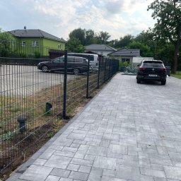 Maja Zäune Aus Polen - Nowoczesne, metalowe ogrodzenie panelowe w kolorze grafitowym, oddzielające posesję z kostki brukowej, z widocznymi dwoma samochodami i zielonym domem w tle.