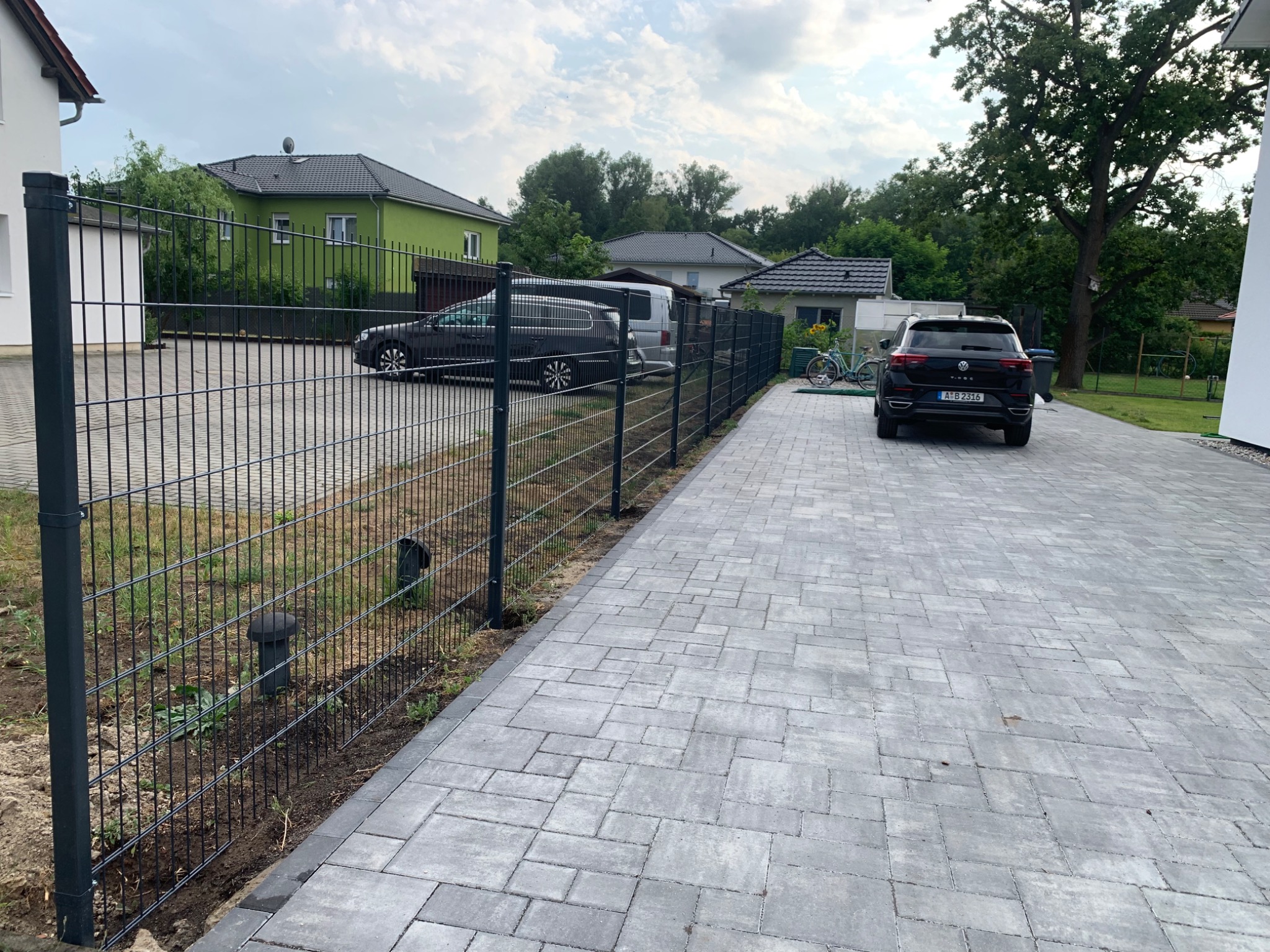 Nowoczesne, metalowe ogrodzenie panelowe w kolorze grafitowym, oddzielające posesję z kostki brukowej, z widocznymi dwoma samochodami i zielonym domem w tle.