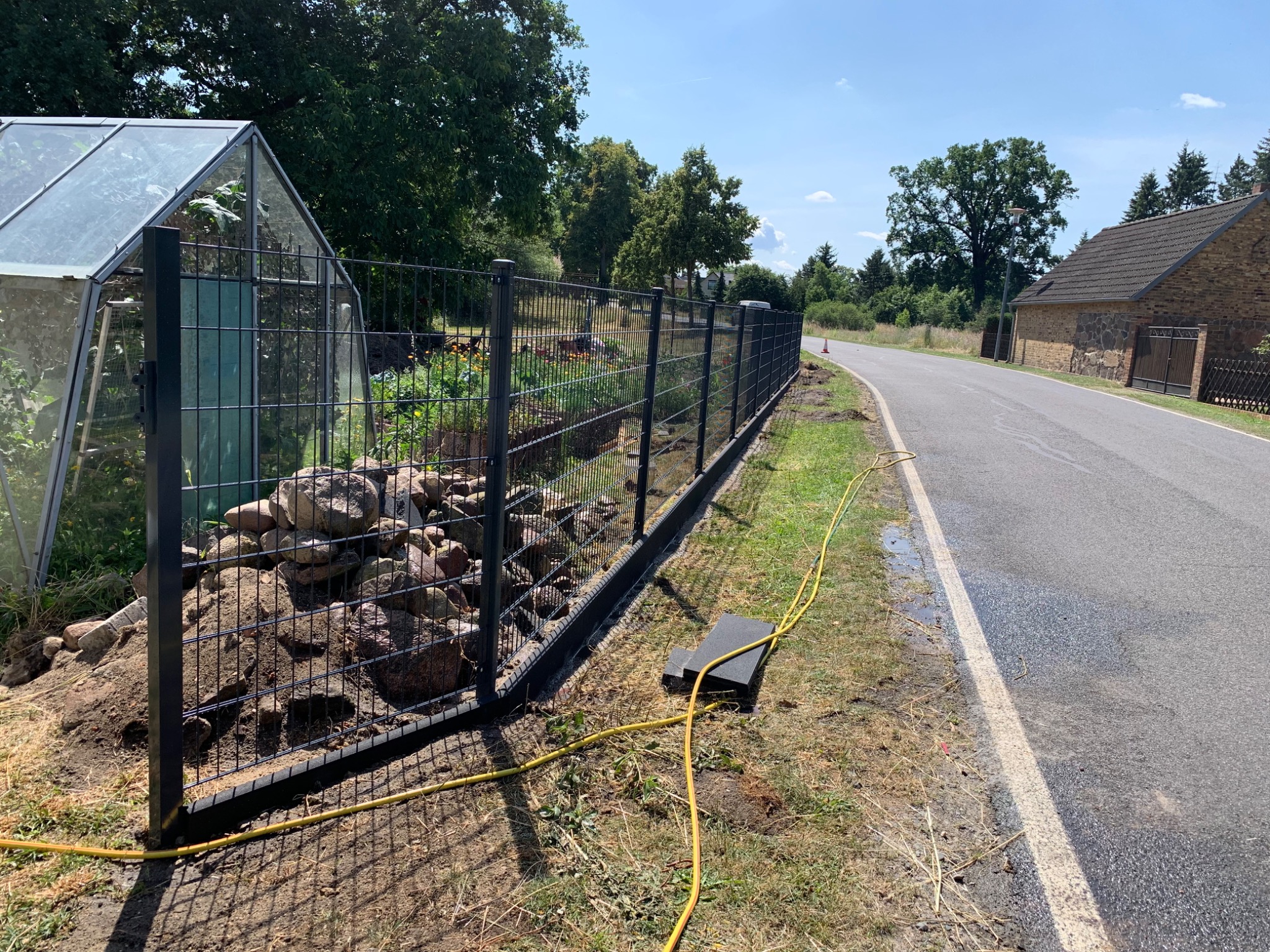 Nowo postawione, czarne, metalowe ogrodzenie panelowe z podmurówką betonową, w tle szklarnia, ziemia i kamienie przygotowane pod nasadzenia, widok z boku na fragment drogi.