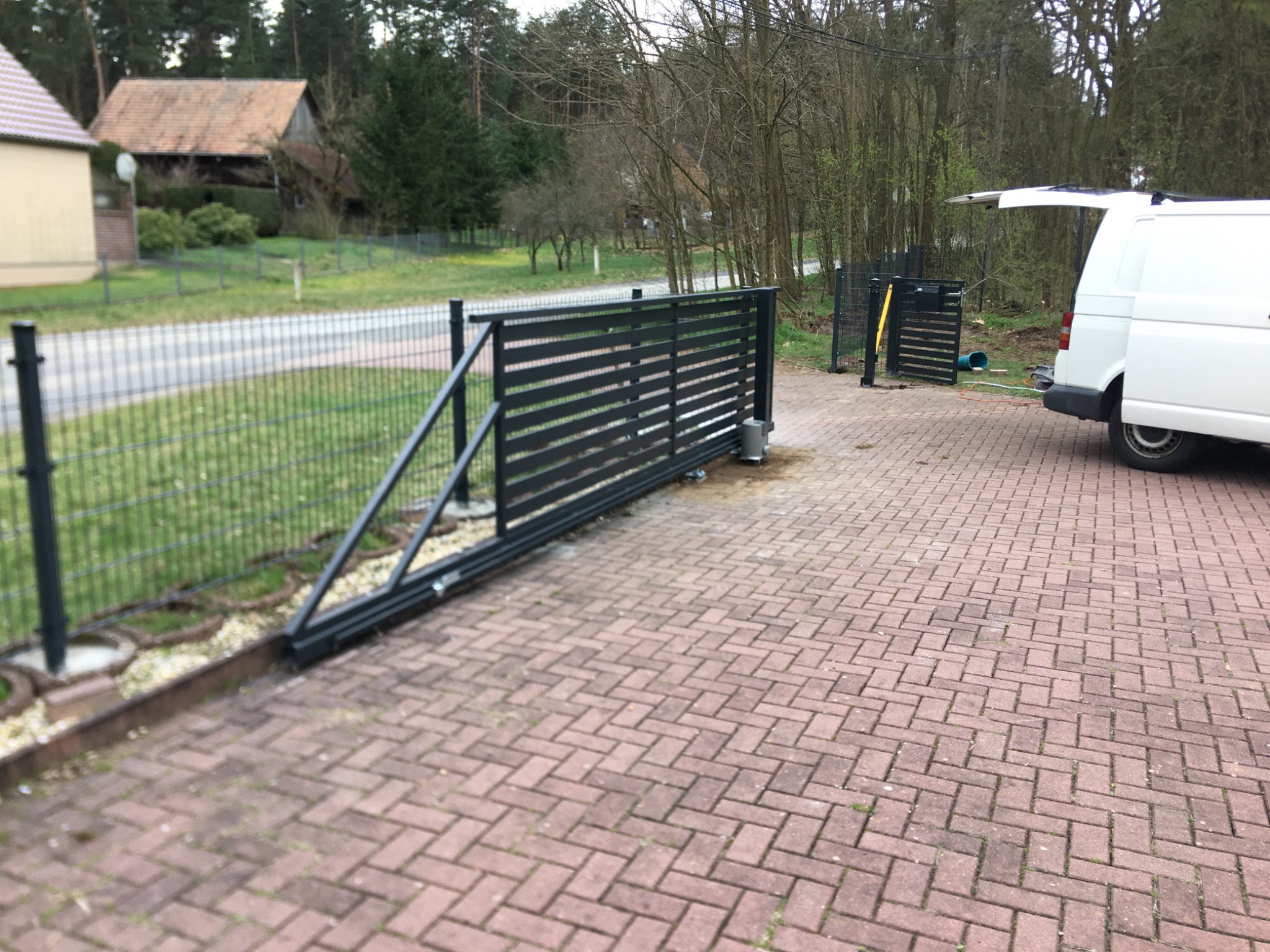 Automatyczna brama przesuwna z poziomymi panelami i napędem, widoczna na tle kostki brukowej i częściowo otwartego białego samochodu dostawczego.