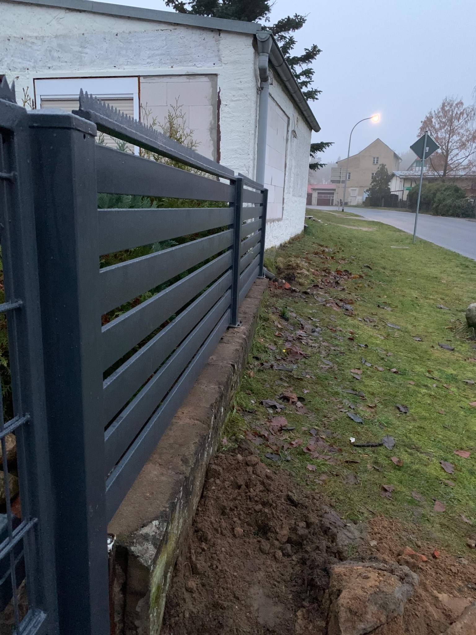 Nowoczesne, poziome ogrodzenie panelowe w kolorze grafitowym z kolcami na górze, zamontowane na betonowej podmurówce, widok na ulicę i budynki w tle w pochmurny dzień.