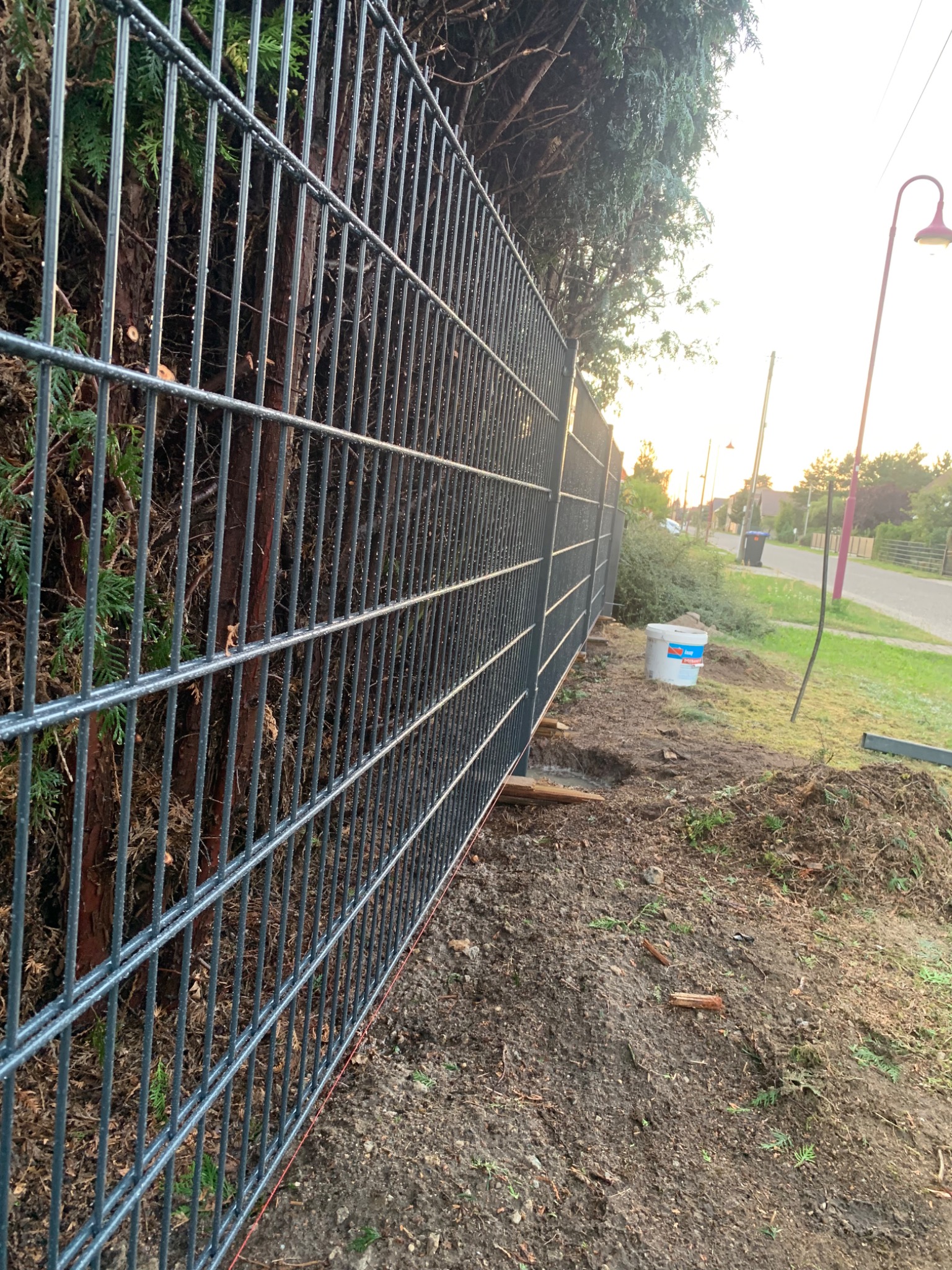 Metalowe ogrodzenie panelowe w trakcie montażu, widoczne wykopane dołki pod słupki, zarośnięte tło z drzewami i elementami krajobrazu.