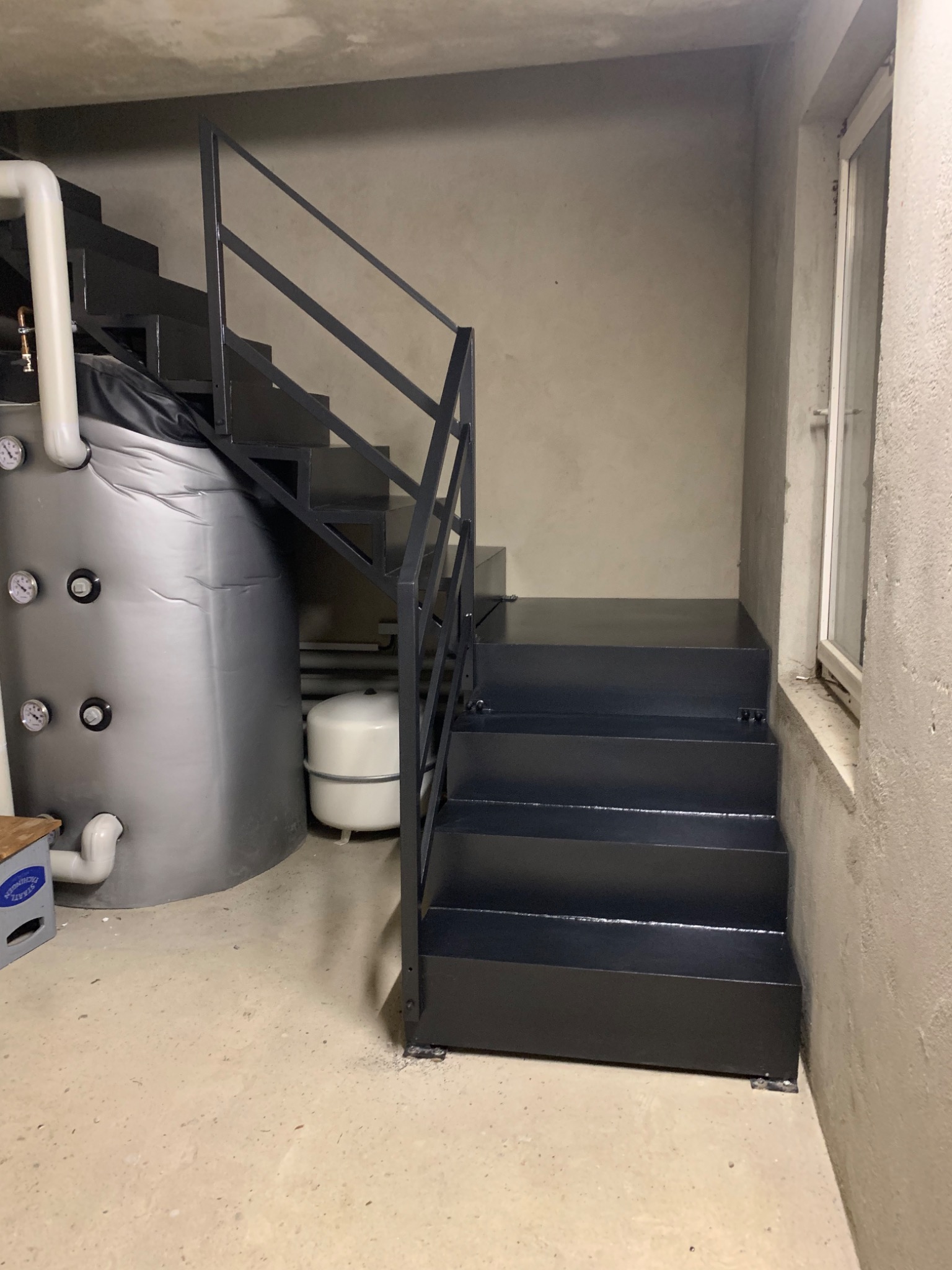 Ciemne, metalowe schody z barierką w szarej piwnicy, obok zbiornik na wodę z manometrami i mały zbiornik wyrównawczy.