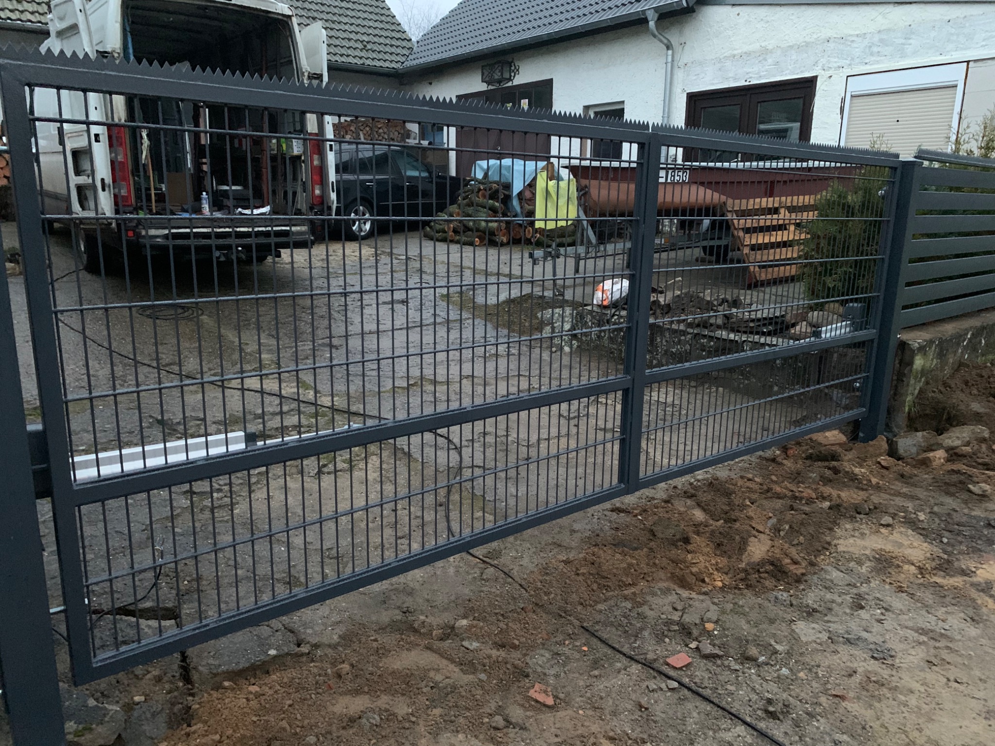 Szara, metalowa brama przesuwna z ostrzami na górze, widok z zewnątrz posesji, w tle zaparkowany biały bus i sterta drewna.
