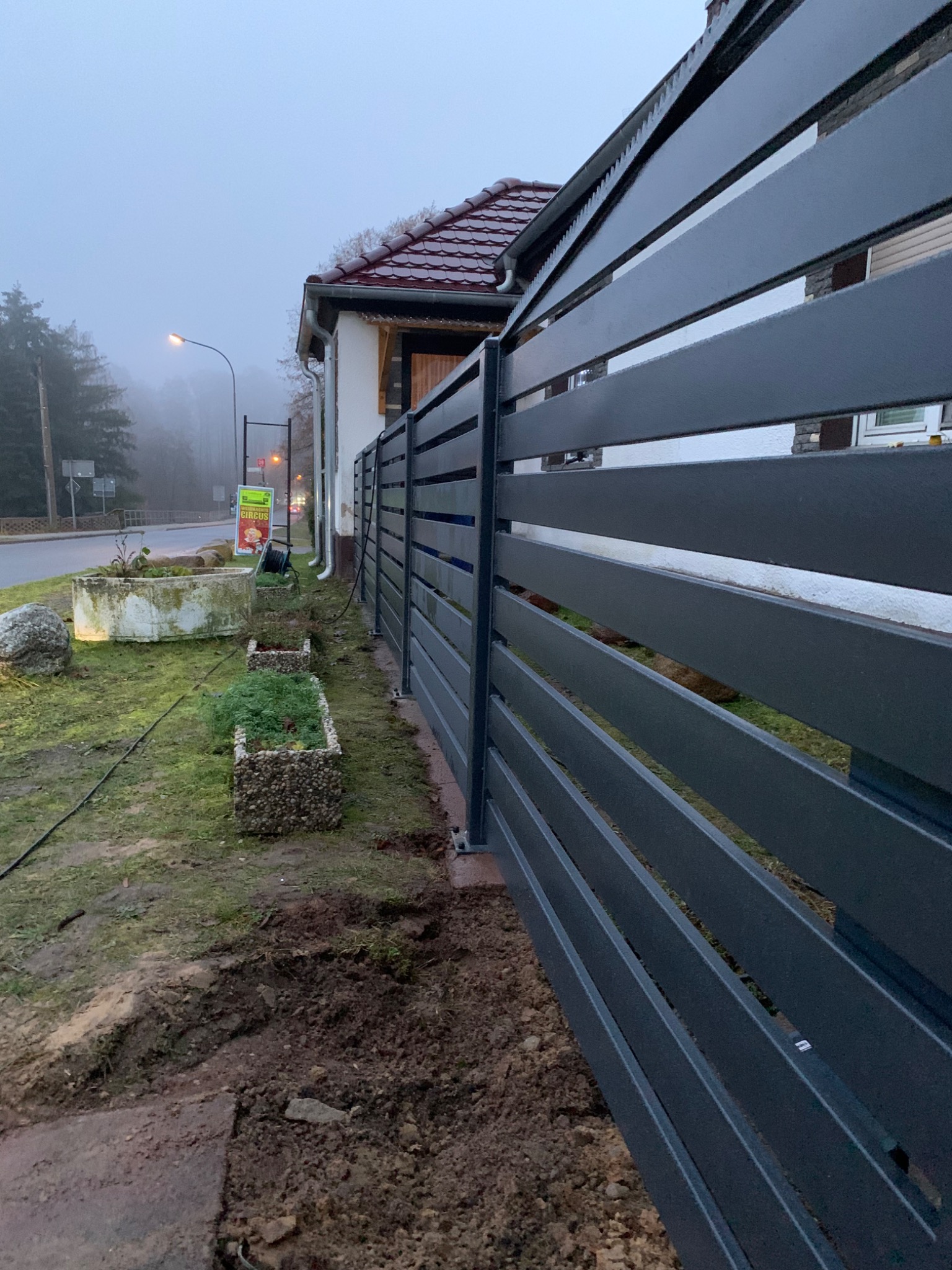 Nowoczesne, poziome ogrodzenie panelowe w kolorze grafitowym, widoczne z bliskiej perspektywy, z ziemią po montażu i elementami zieleni w tle, w pochmurny dzień.