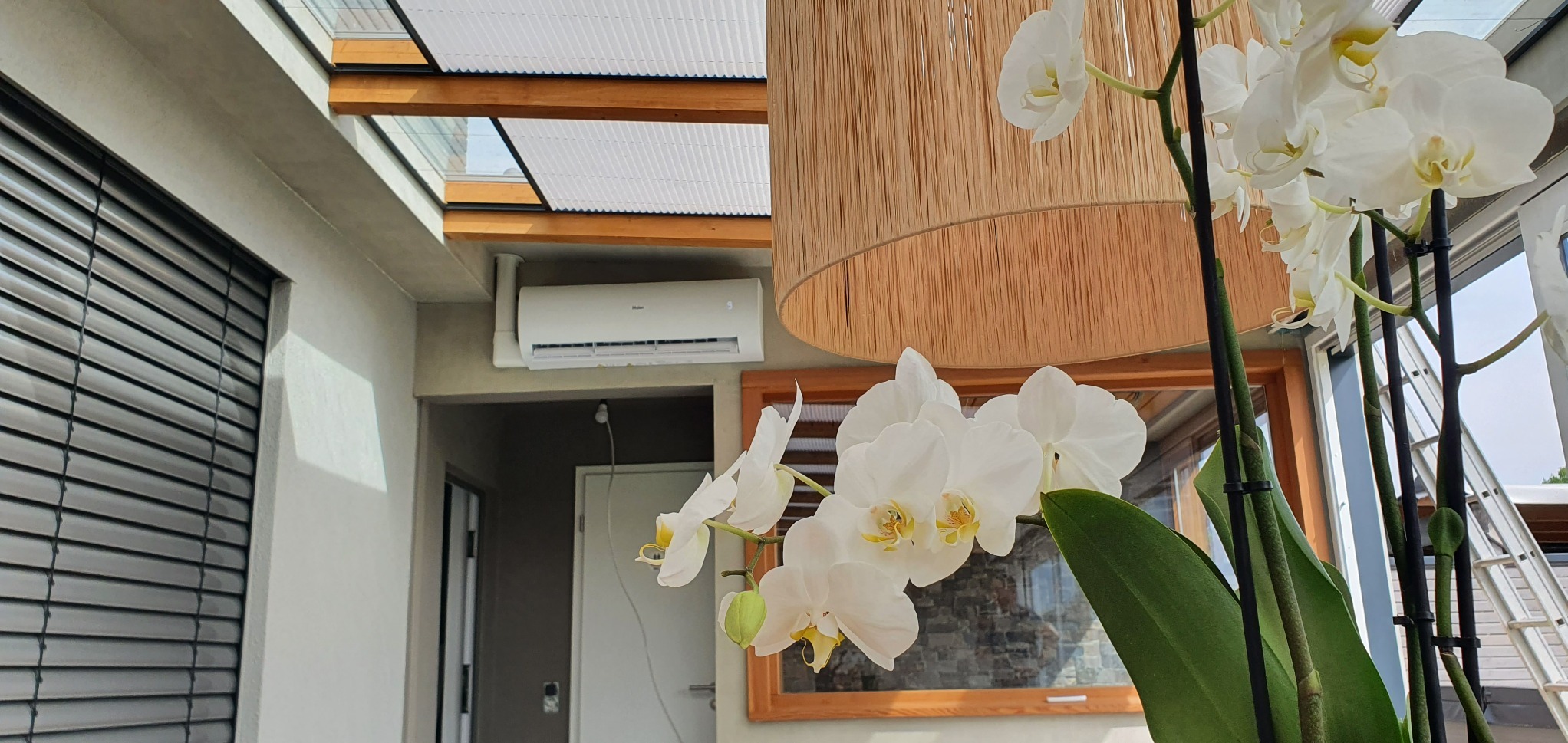 Biała klimatyzacja Haier na ścianie w nowoczesnym wnętrzu z przeszklonym dachem, widoczna przez kwiaty orchidei i abażur lampy.