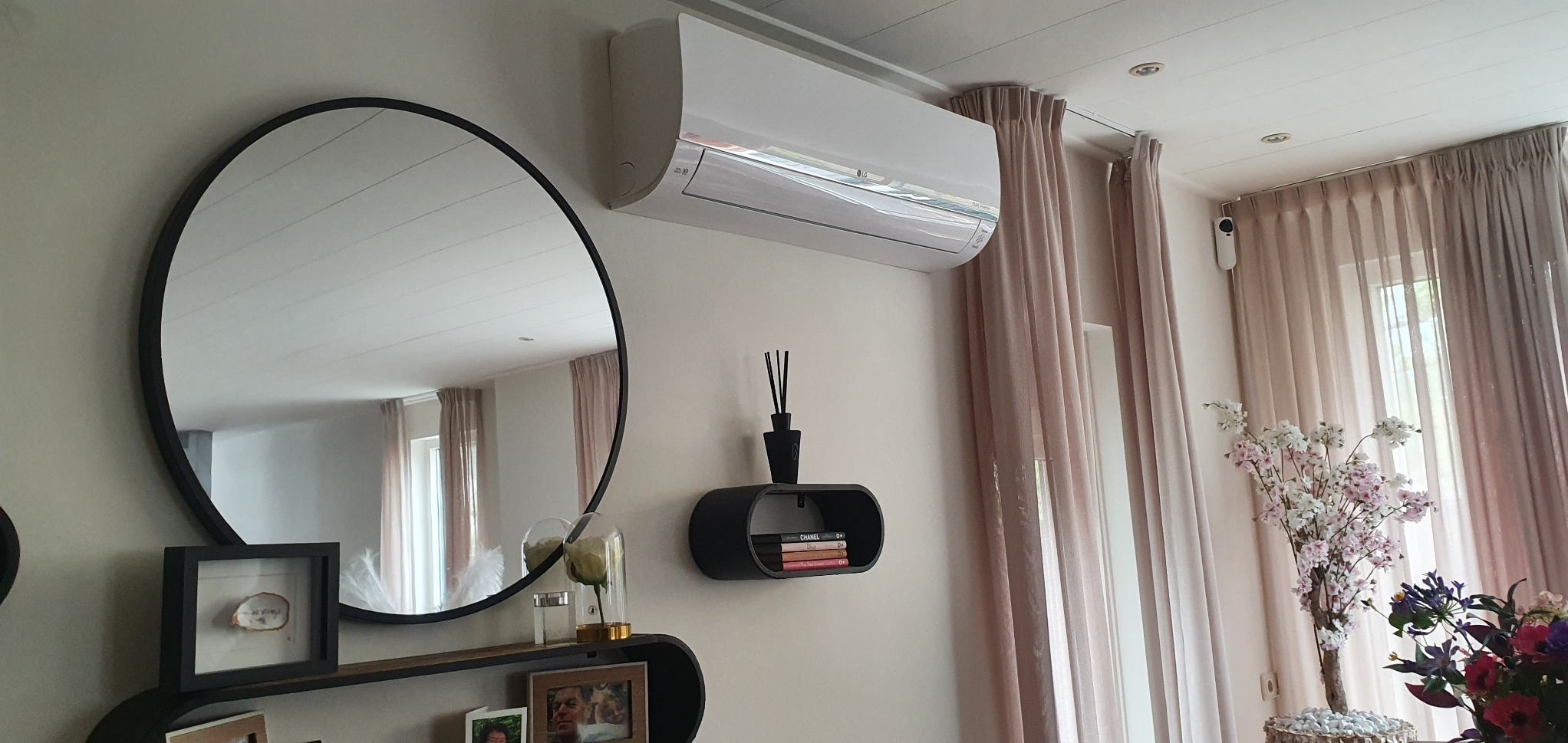 Nowoczesny klimatyzator LG na ścianie w salonie, obok okrągłe lustro i dekoracje. Jasne wnętrze z beżowymi zasłonami i kwiatami.