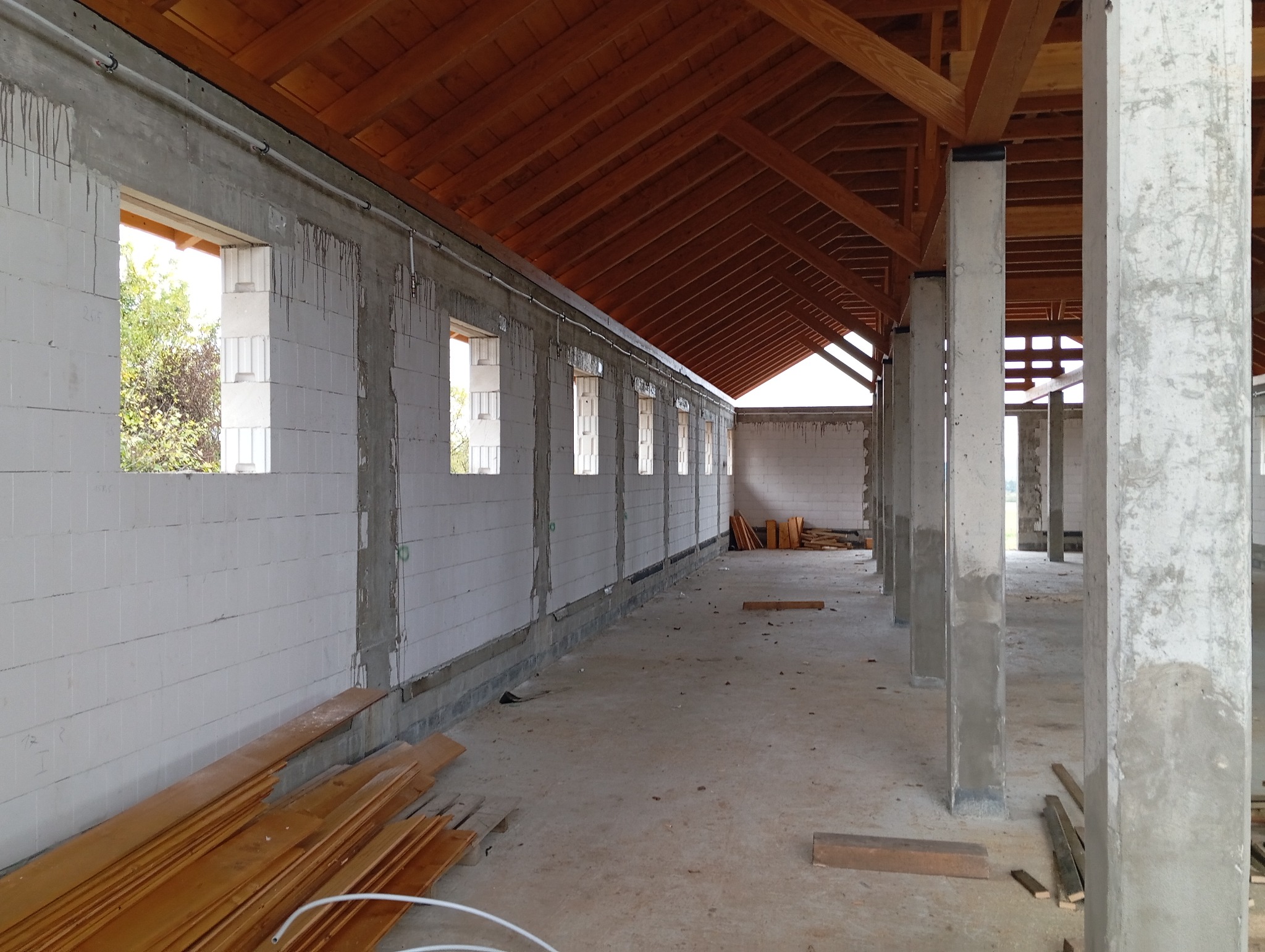 Wnętrze surowego budynku w trakcie budowy, widoczne ściany z białych bloczków, betonowe słupy i drewniana konstrukcja dachu, z widocznymi przewodami instalacji pod sufitem.