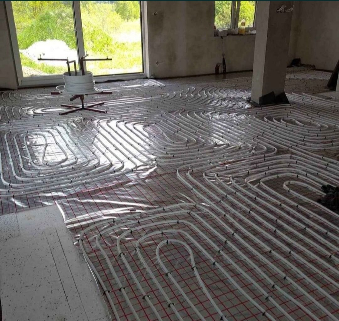 Białe rury instalacji ogrzewania podłogowego rozłożone na czerwonej siatce w surowym wnętrzu z oknem.