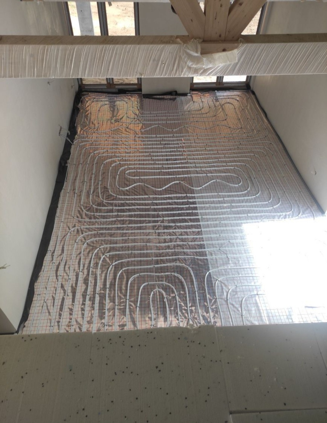 Instalacja ogrzewania podłogowego: białe rurki ułożone w pętle na srebrnej folii izolacyjnej z siatką, widok z góry na surowy stan podłoża.
