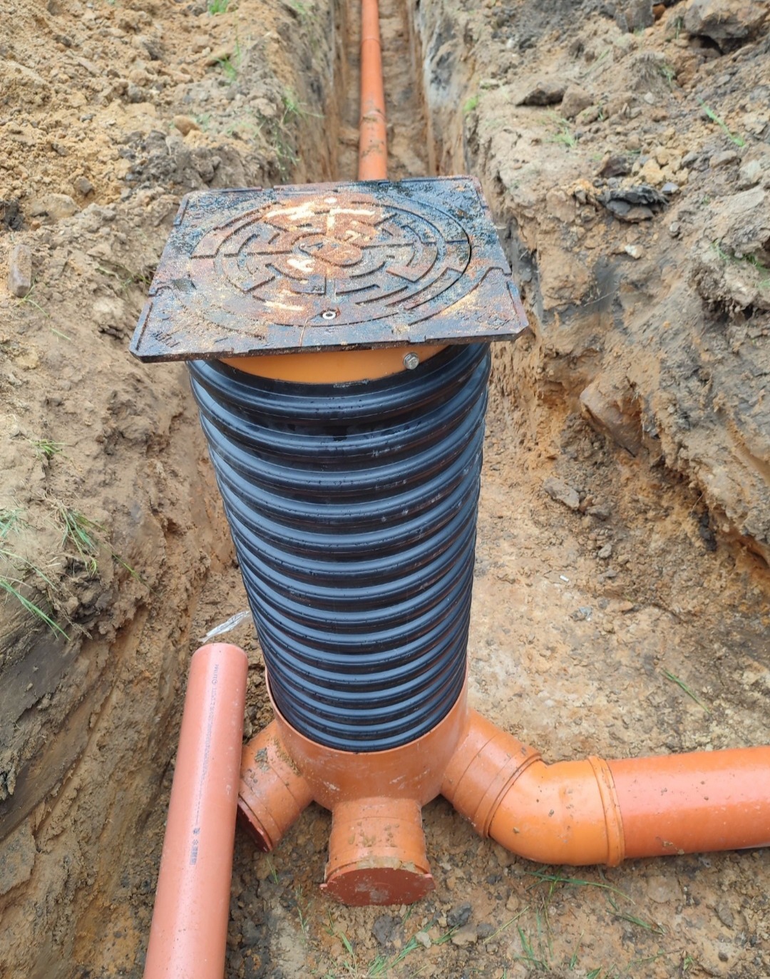 Studzienka kanalizacyjna z rurami w wykopie, widoczna pokrywa z żeliwa i karbowana rura pionowa.