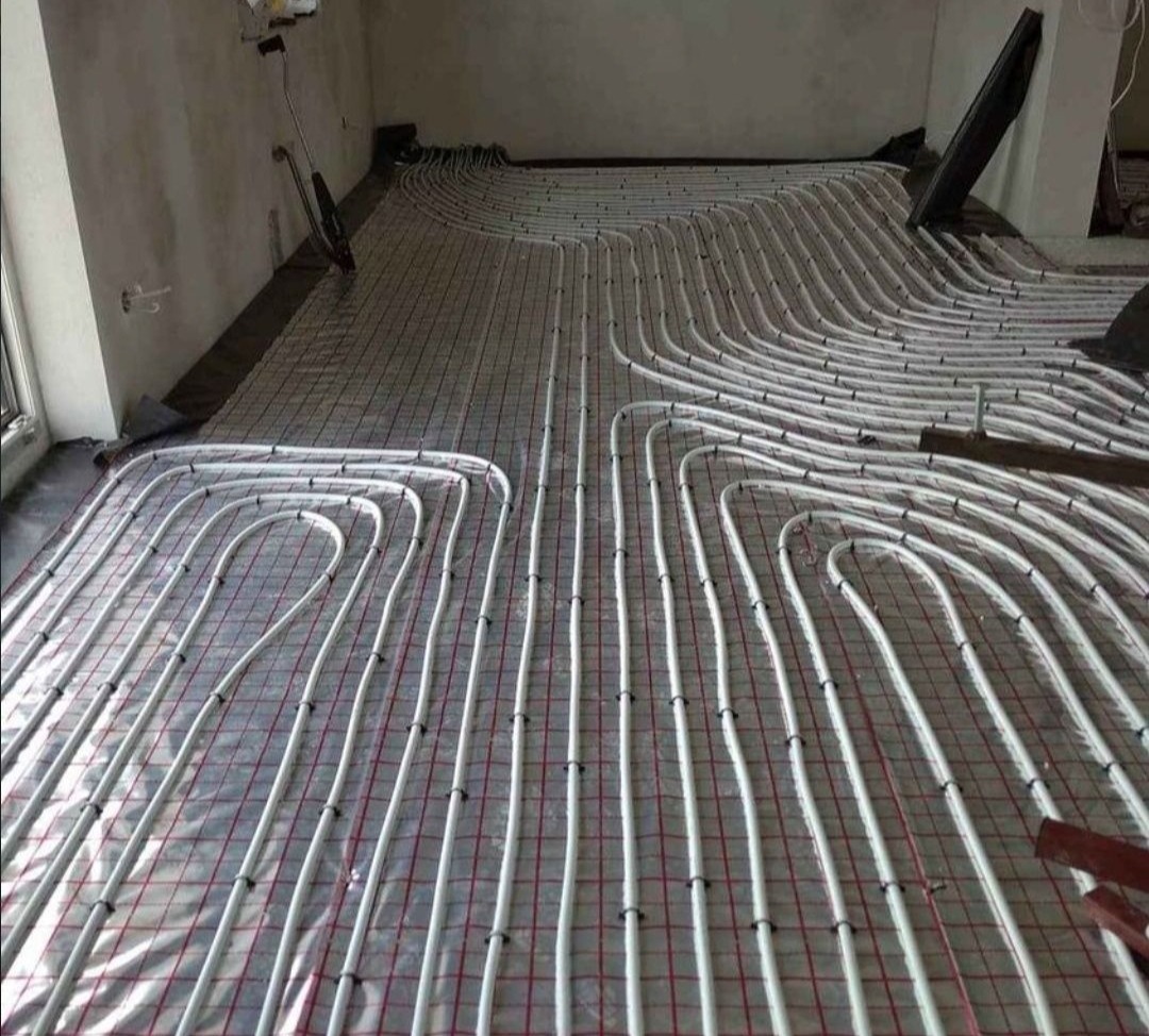 Białe rury ogrzewania podłogowego ułożone na siatce w surowym wnętrzu przed wylewką.