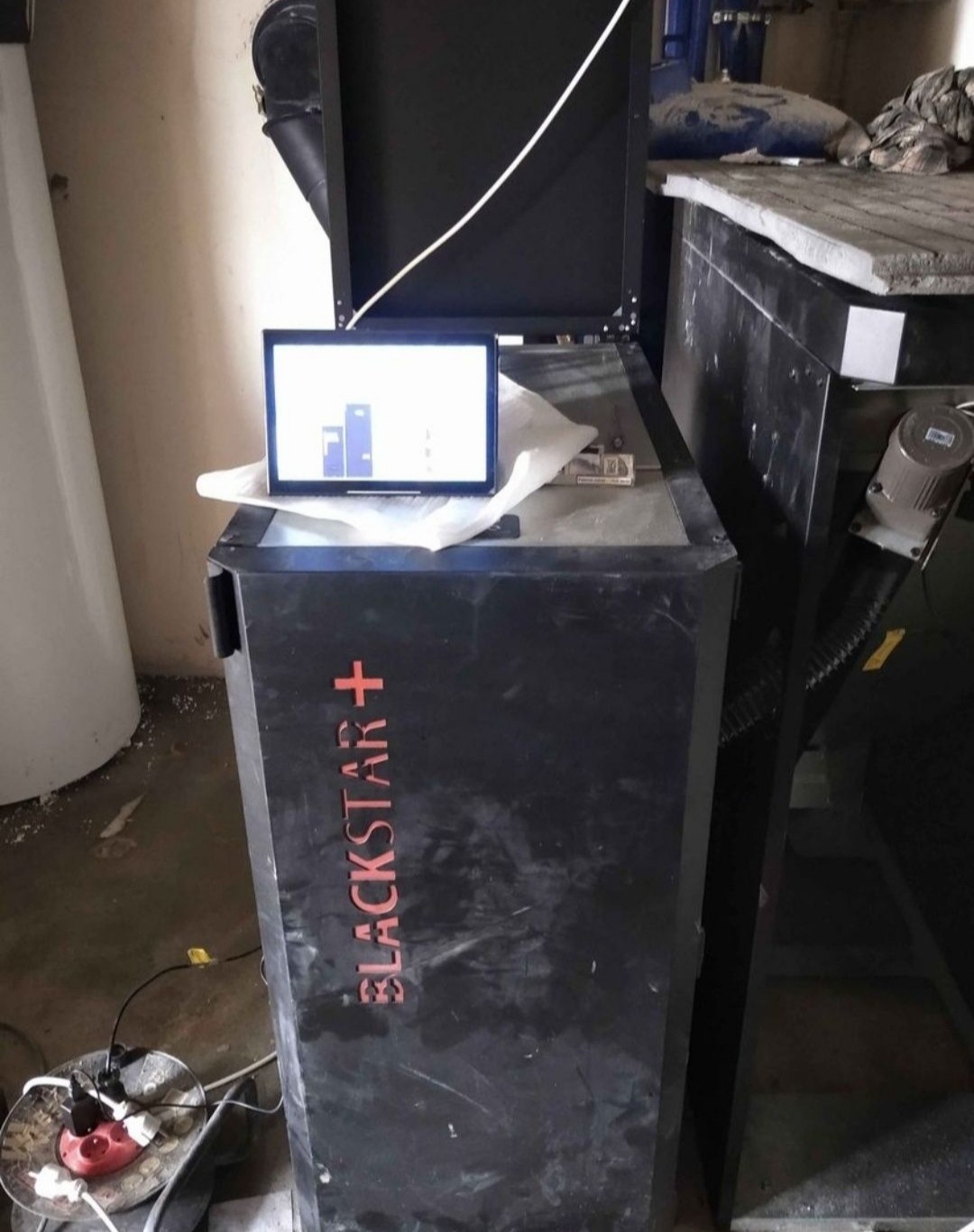 Czarny piec z wyświetlaczem pokazującym wykres słupkowy, model Blackstar+, widoczny kabel zasilający i otoczenie kotłowni.