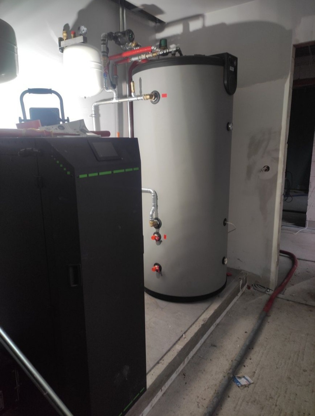 Szary zasobnik ciepłej wody użytkowej z podłączonymi rurami i zaworami, stojący obok czarnego kotła na paliwo stałe w surowym, betonowym pomieszczeniu.