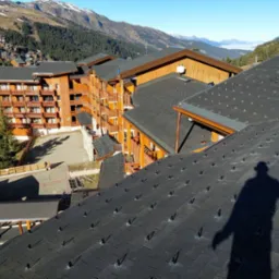 Nowy dach z czarnej dach&oacute;wki bitumicznej na budynku w g&oacute;rskim kurorcie, widoczny cień osoby fotografującej, w tle zabudowania i ośnieżone szczyty.