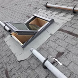 Otwarty świetlik w dachu pokrytym szarym gontem bitumicznym, widoczne rury odgromowe i ślady po naprawach na papie wokół świetlika.