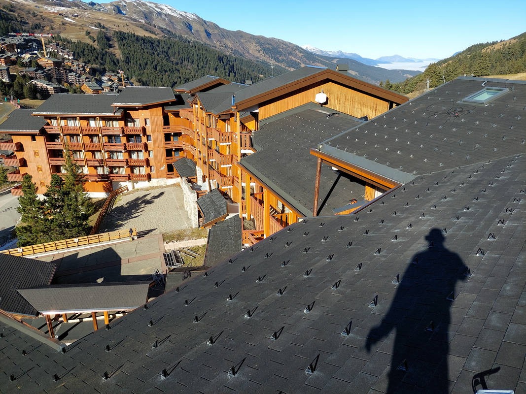 Nowy dach z czarnej dachówki bitumicznej na budynku w górskim kurorcie, widoczny cień osoby fotografującej, w tle zabudowania i ośnieżone szczyty.