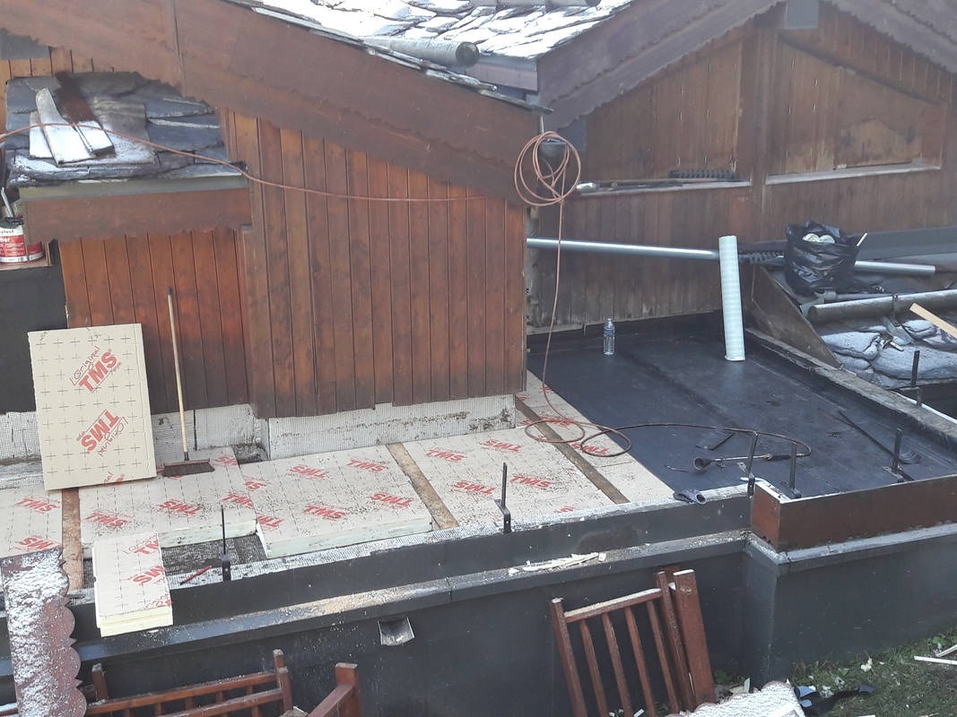 Fragment dachu z drewnianą boazerią, na którym układane są płyty izolacyjne TMS. Widoczne narzędzia dekarskie i częściowo pokryty papą dach. W tle stara dachówka.