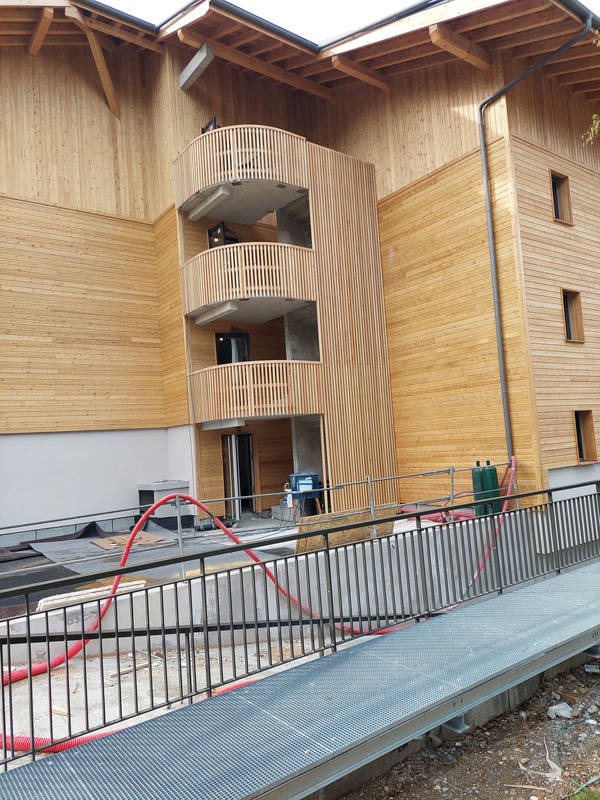 Budynek w trakcie budowy z drewnianą elewacją i trzema balkonami z drewnianymi balustradami, widoczny pomost z metalową kratą i czerwony wąż na placu budowy.