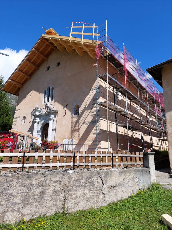 Budynek z odsłoniętą więźbą dachową, zabezpieczony rusztowaniem i siatką ochronną, widoczne detale architektoniczne, ogrodzenie i zieleń w otoczeniu.