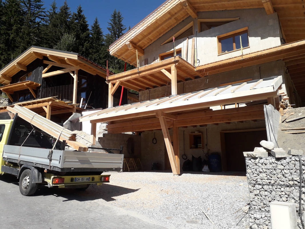 Dwa domy w trakcie budowy, widoczna konstrukcja drewniana dachu, jeden dom z szarą elewacją, drugi z czarną, na pierwszym planie żółta ciężarówka z drewnem.