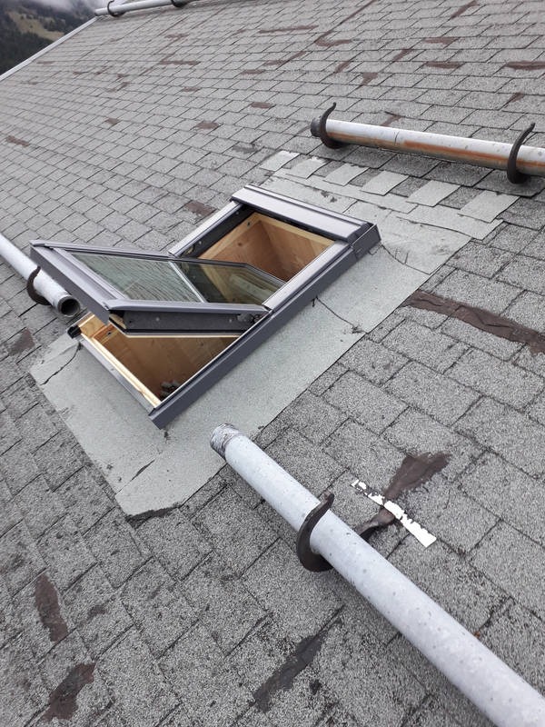 Otwarty świetlik w dachu pokrytym szarym gontem bitumicznym, widoczne rury odgromowe i ślady po naprawach na papie wokół świetlika.