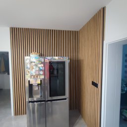 Aleksandra Szempruch - Ściana wykończona drewnianymi lamelami w salonie, za srebrną lodówką z magnesami, widok na narożnik pomieszczenia.