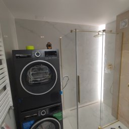 Aleksandra Szempruch - Nowoczesna łazienka z pralko-suszarką Bosch w kolorze grafitowym i kabiną prysznicową ze złotymi profilami, marmurowe płytki na ścianach, widoczny poziomica laserowa.