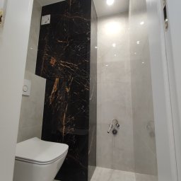 Aleksandra Szempruch - Wykończona łazienka w Kraśniku z toaletą podwieszaną i ścianą z czarnego marmuru z złotymi żyłami, widoczne płytki w odcieniach szarości oraz białe drzwi.