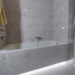 Aleksandra Szempruch - Wykończona łazienka z wanną obłożoną płytkami imitującymi marmur i mozaiką, z podświetleniem LED u dołu, oraz grzejnikiem drabinkowym z żółtym ręcznikiem.