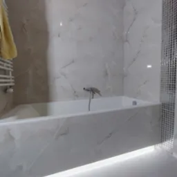 Wykończona łazienka z wanną obłożoną płytkami imitującymi marmur i mozaiką, z podświetleniem LED u dołu, oraz grzejnikiem drabinkowym z żółtym ręcznikiem.