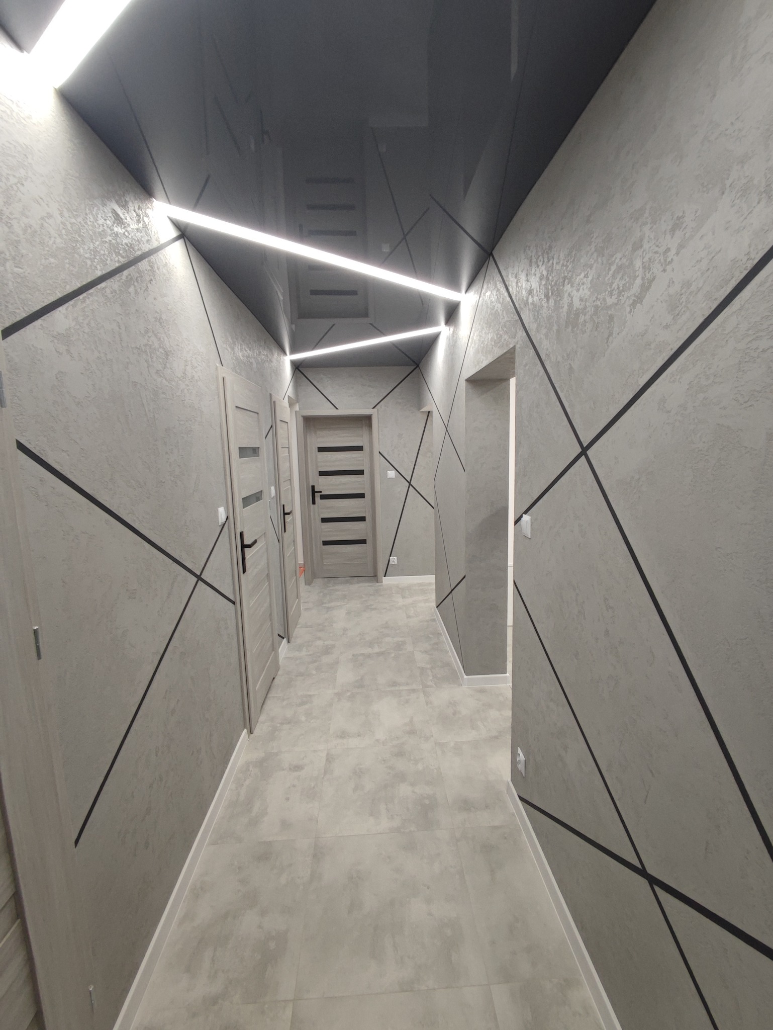 Korytarz po remoncie w odcieniach szarości z gładzią na ścianach, czarnymi geometrycznymi wzorami, podwieszanym, lustrzanym sufitem z ledowym oświetleniem i trzema parami drzwi. Podłoga wyłożona...