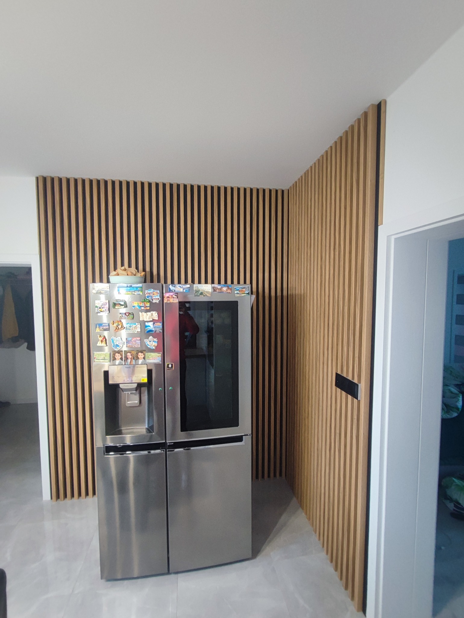 Ściana wykończona drewnianymi lamelami w salonie, za srebrną lodówką z magnesami, widok na narożnik pomieszczenia.