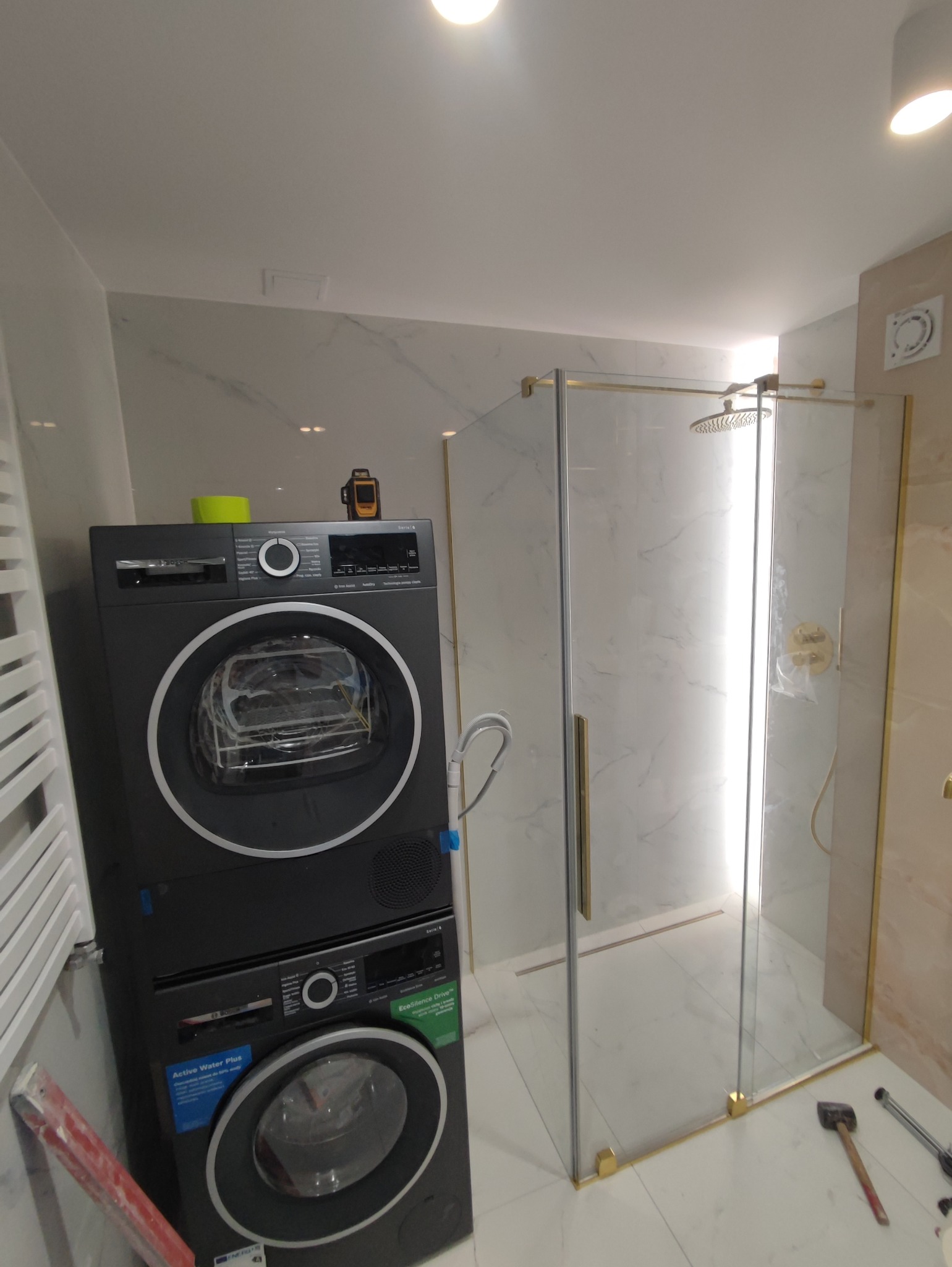 Nowoczesna łazienka z pralko-suszarką Bosch w kolorze grafitowym i kabiną prysznicową ze złotymi profilami, marmurowe płytki na ścianach, widoczny poziomica laserowa.