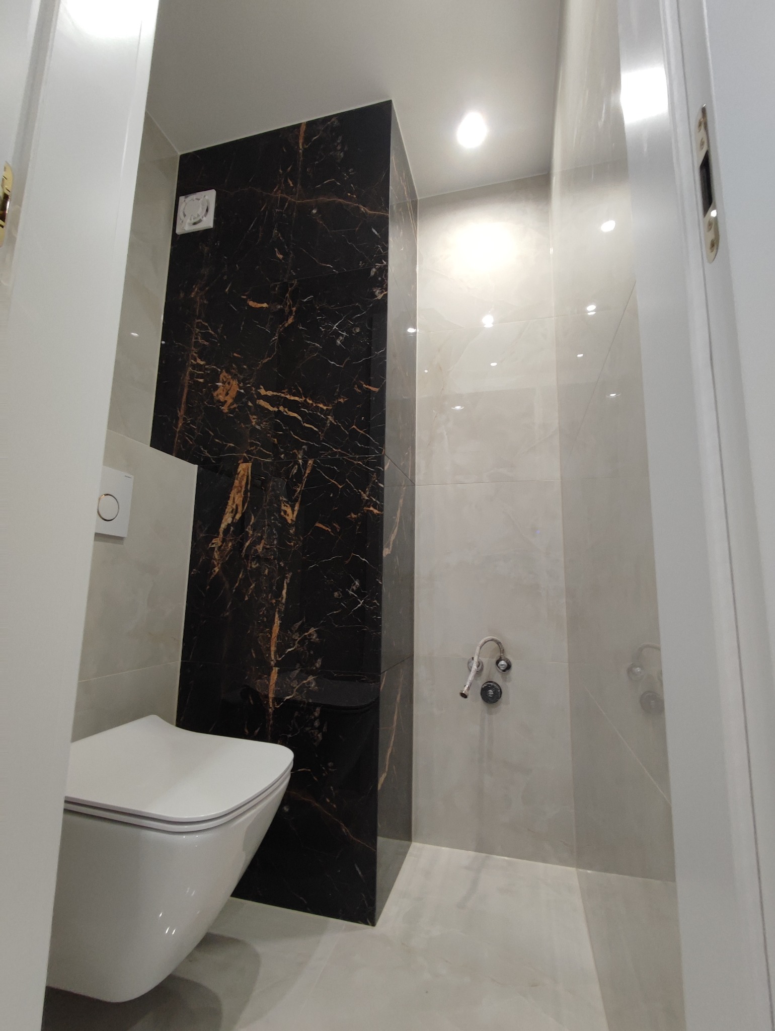 Wykończona łazienka w Kraśniku z toaletą podwieszaną i ścianą z czarnego marmuru z złotymi żyłami, widoczne płytki w odcieniach szarości oraz białe drzwi.