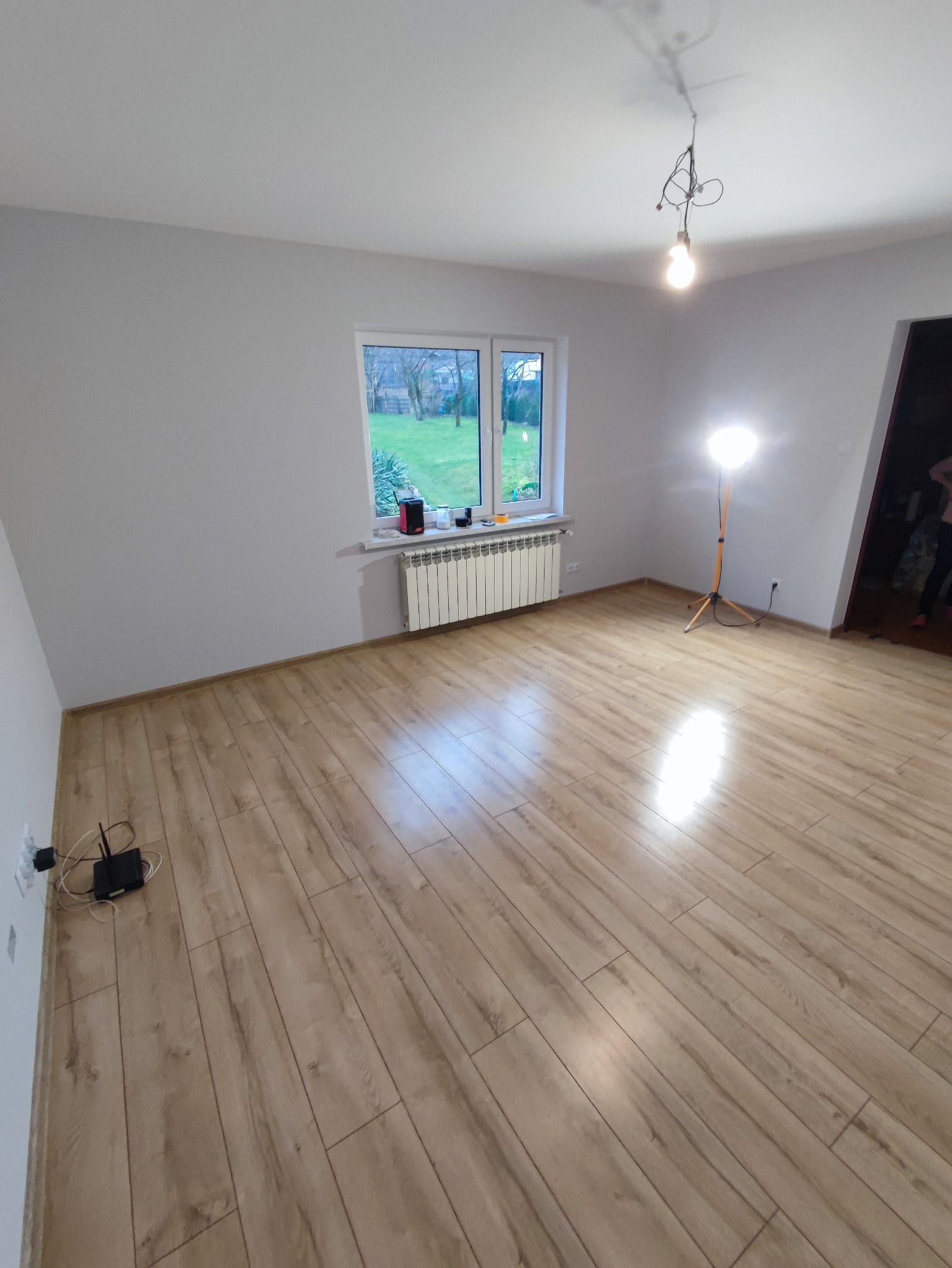 Pusty pokój po położeniu paneli podłogowych, widoczny grzejnik pod oknem, lampa budowlana i wystające kable od routera.