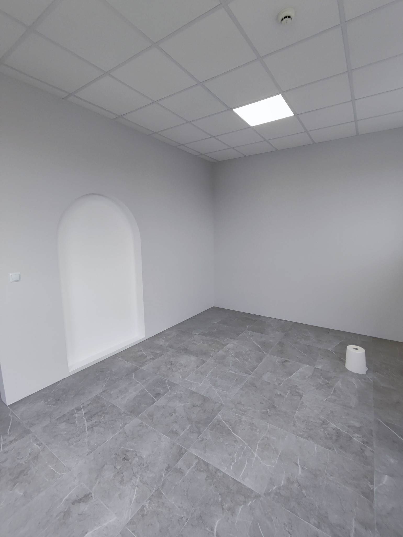 Wnętrze pokoju z szarą marmurową podłogą, białe ściany z niszą w kształcie łuku, sufit kasetonowy z kwadratową lampą i rolką papieru toaletowego w rogu.