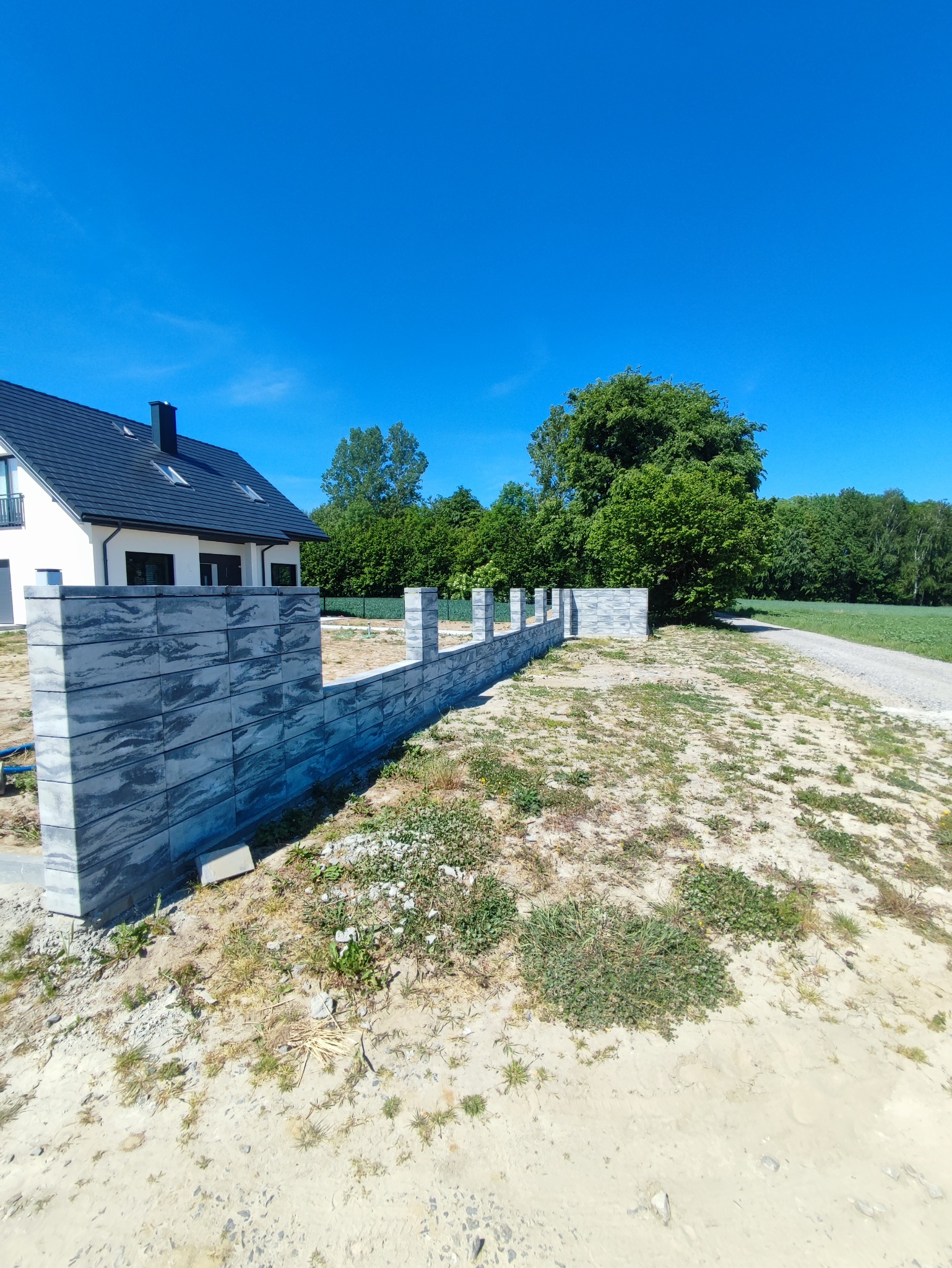 Wznoszony mur z szarych, betonowych bloczków o falistej fakturze, oddzielający posesję z nowoczesnym domem od polnej drogi w słoneczny dzień.