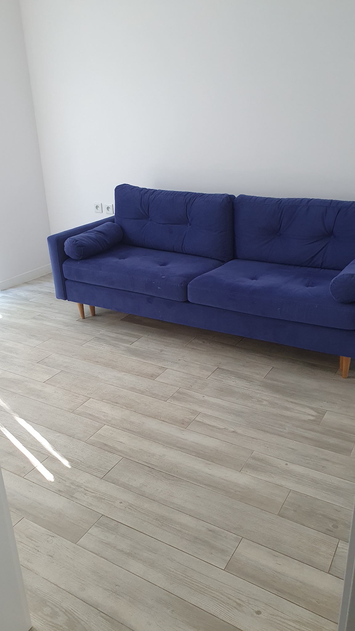 Nowoczesna, granatowa sofa z poduszkami na drewnianych nóżkach, ustawiona na podłodze z paneli imitujących drewno, na tle białej ściany z gniazdkami elektrycznymi.