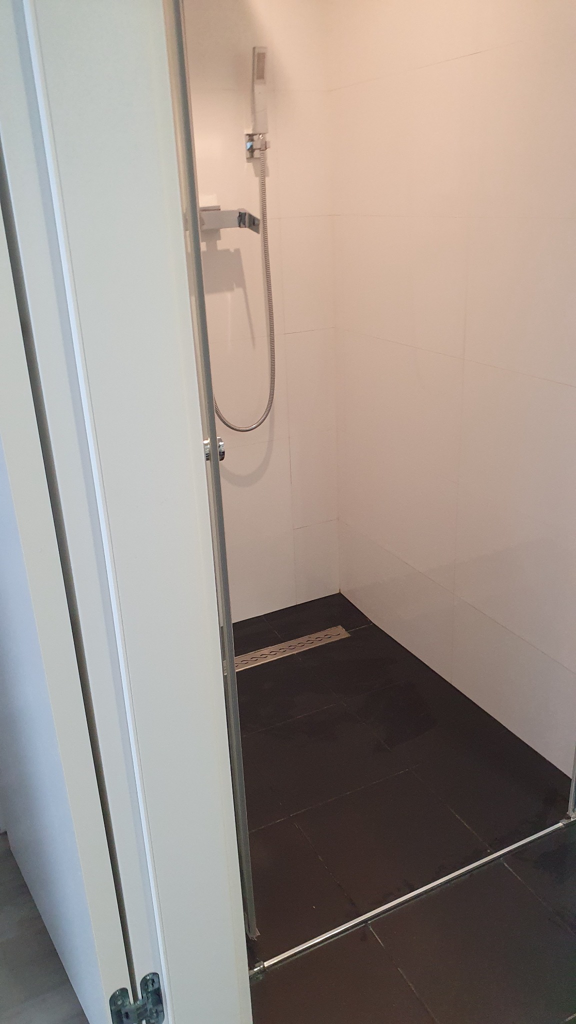 Nowoczesna kabina prysznicowa z czarną podłogą i odpływem liniowym, widok z zewnątrz przez uchylone białe drzwi.