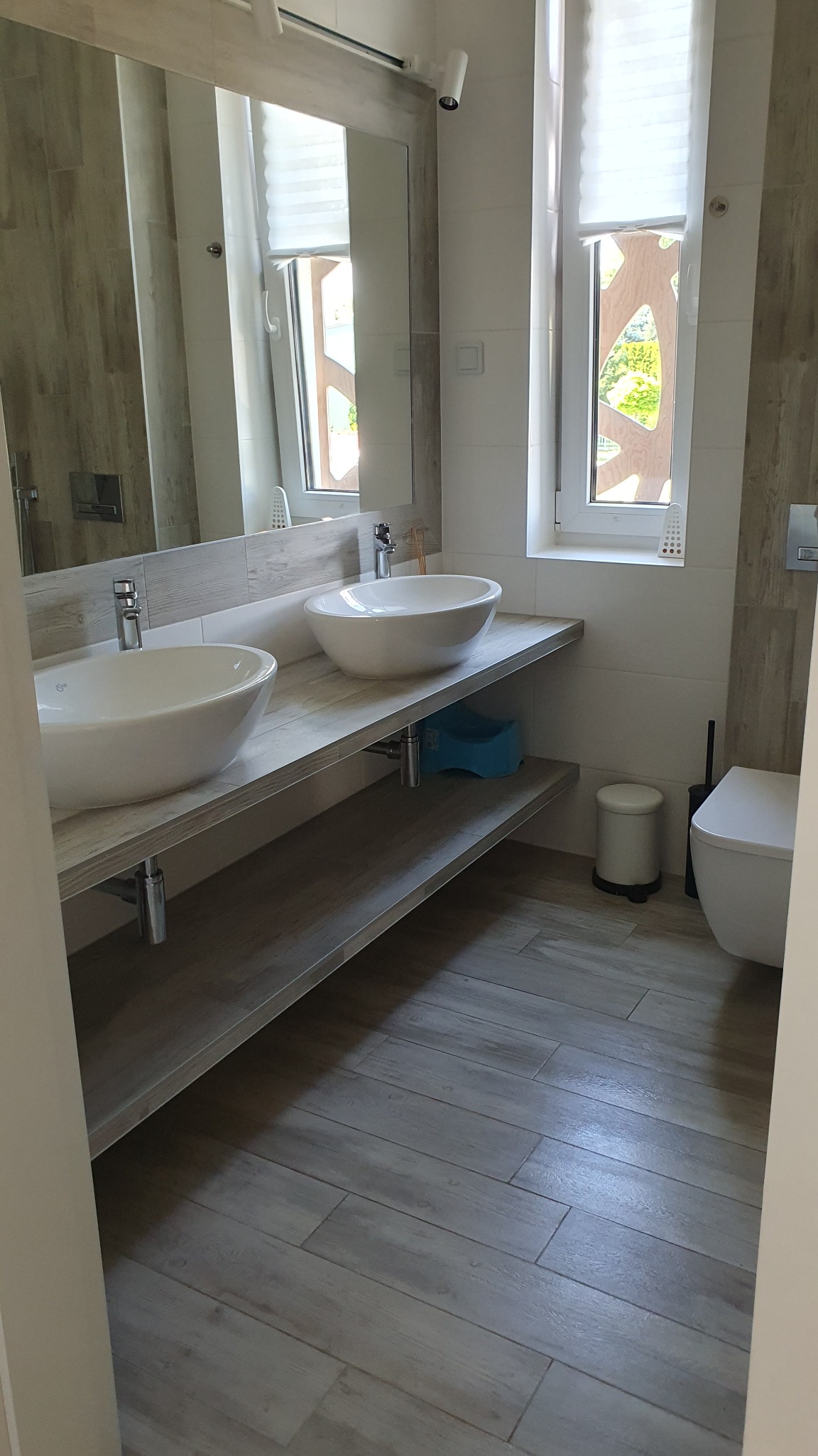 Jasna łazienka z dwoma okrągłymi umywalkami nablatowymi, drewnopodobnymi płytkami na podłodze i ścianach, oraz oknem z żaluzjami plisowanymi.