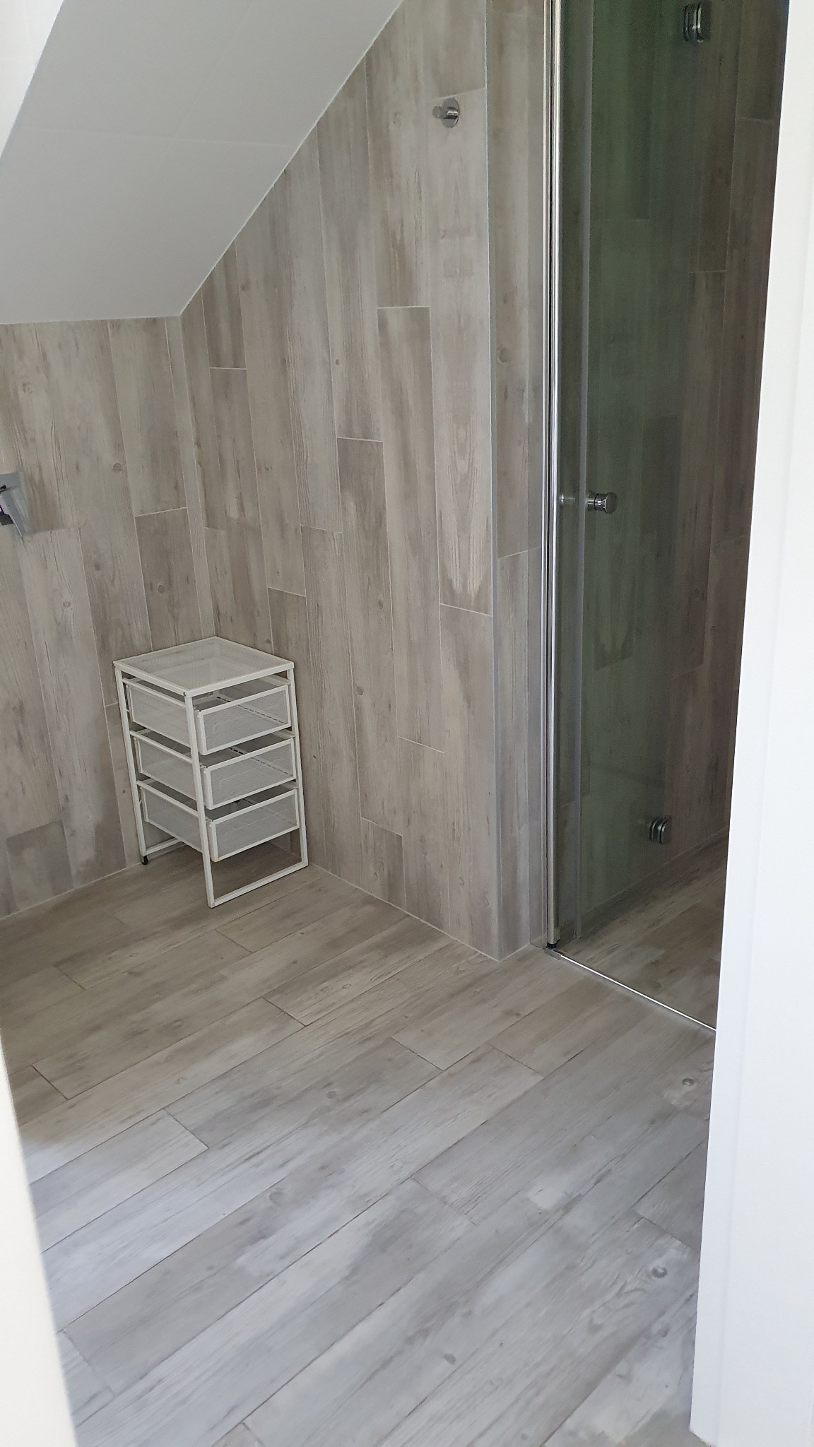 Jasna łazienka z kabiną prysznicową i białym regałem na tle drewnopodobnych płytek ściennych i podłogowych.