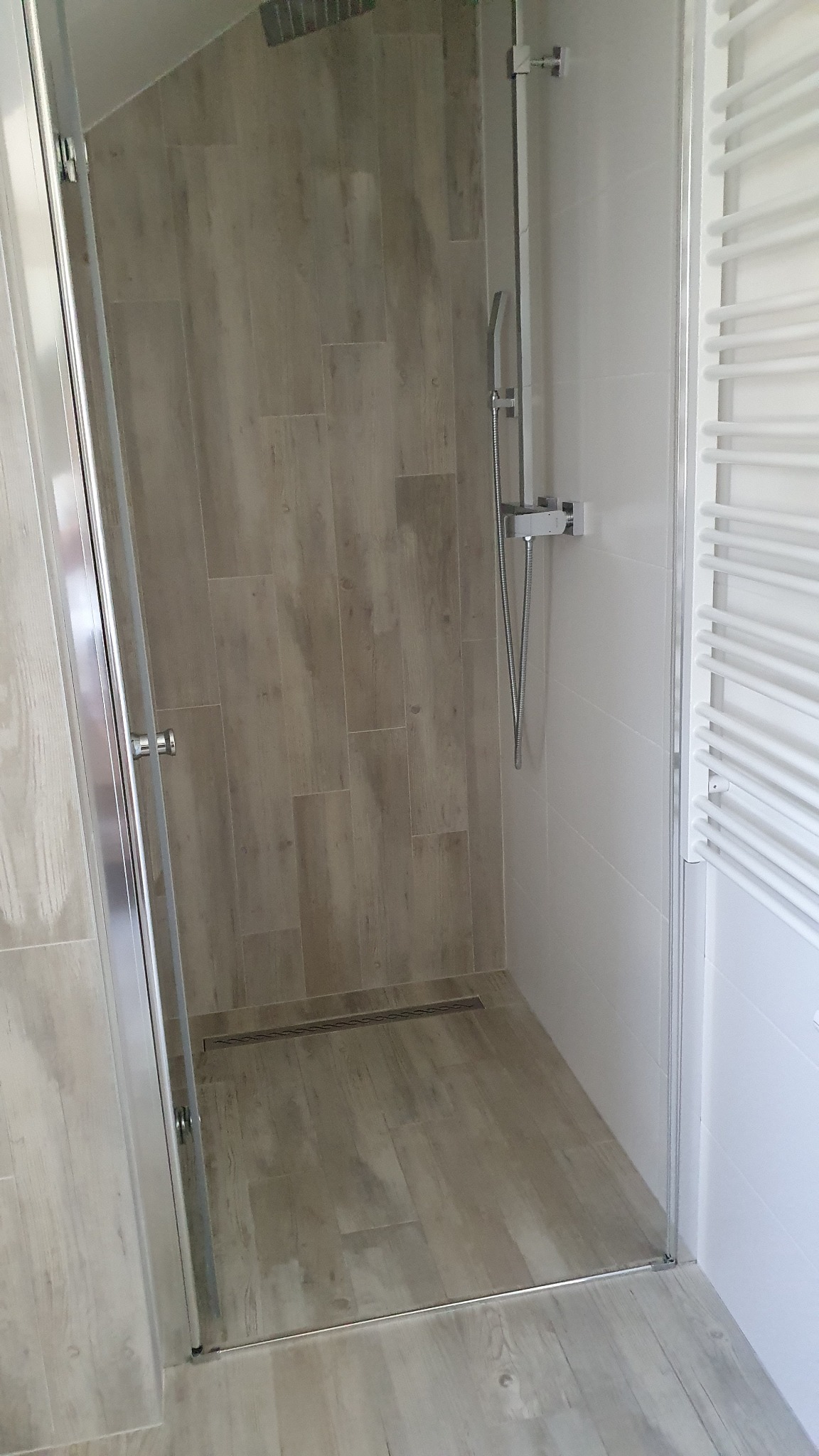 Kabina prysznicowa z odpływem liniowym w podłodze wyłożonej płytkami imitującymi drewno, chromowana bateria i deszczownica, grzejnik drabinkowy.