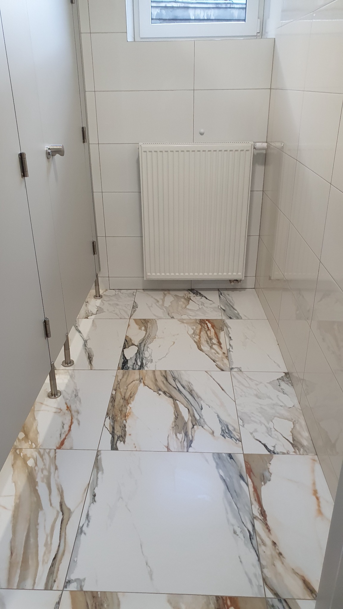 Wnętrze publicznej toalety z białymi kabinami, grzejnikiem i podłogą z płytek imitujących marmur.