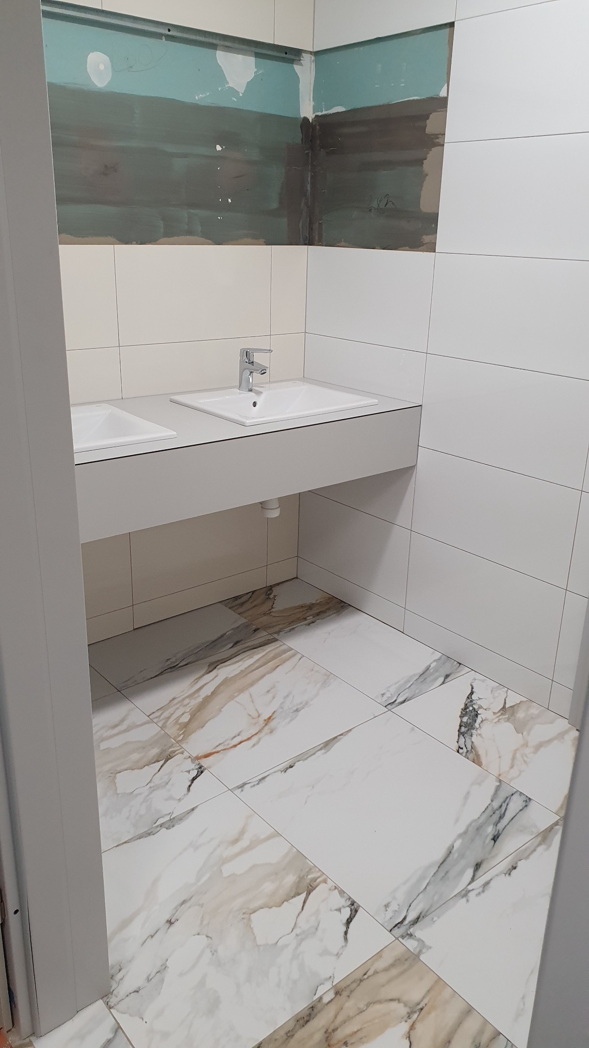 Wnętrze łazienki w trakcie remontu, widoczne dwie umywalki z bateriami, białe płytki na ścianach oraz marmurowe płytki na podłodze, ściana w górnej części nieotynkowana.