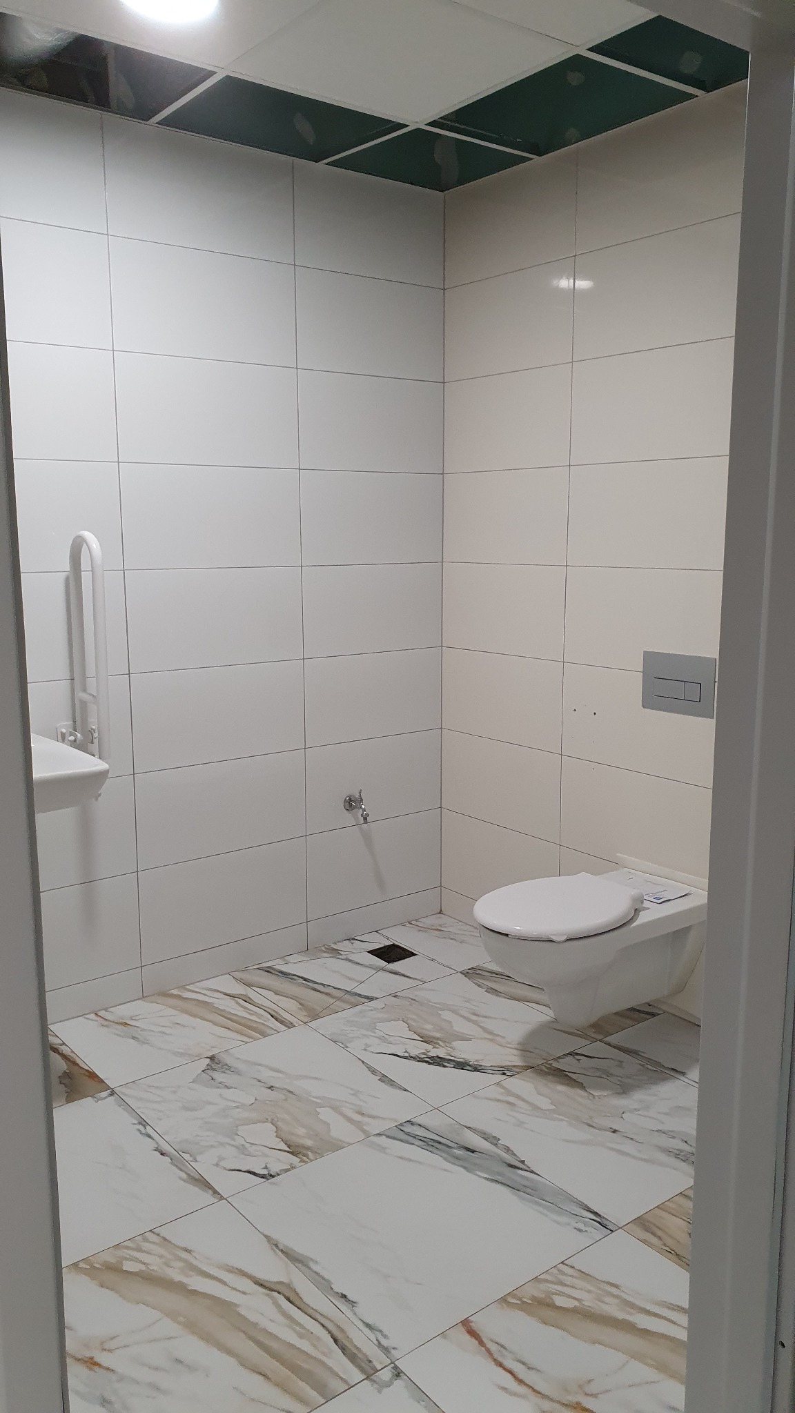 Wykończona łazienka z białymi płytkami ściennymi, marmurową podłogą, umywalką z uchwytem dla niepełnosprawnych i podwieszaną toaletą, widok zza futryny drzwi.