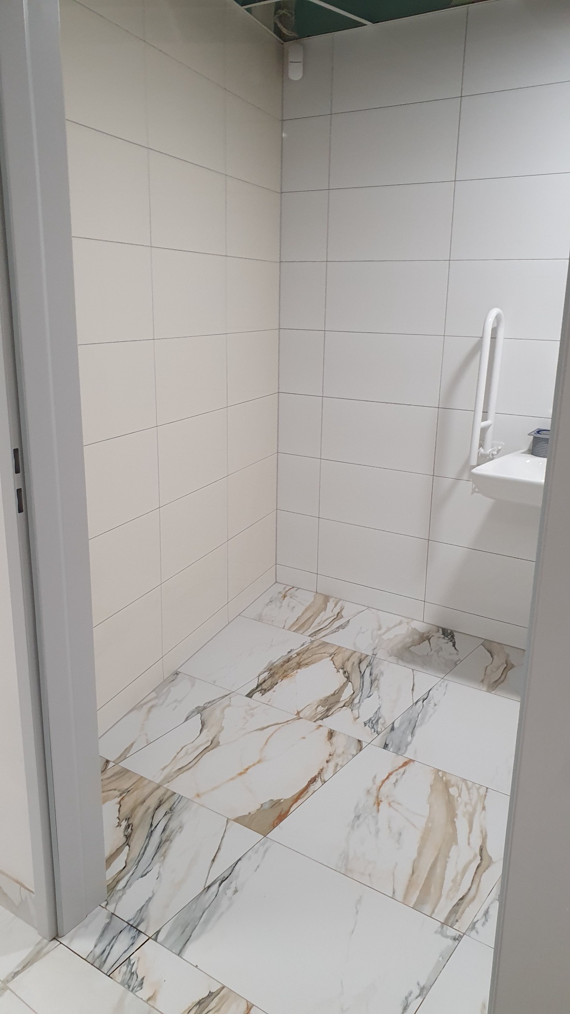 Wnętrze łazienki z białymi płytkami ściennymi i podłogą imitującą marmur, widoczna umywalka z uchwytem dla niepełnosprawnych oraz fragment sufitu podwieszanego.