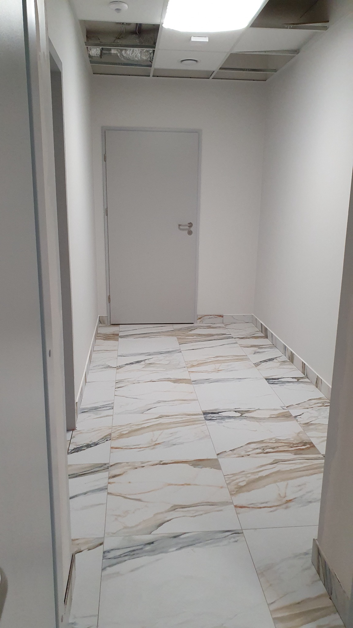 Korytarz biurowy z marmurową podłogą, białą ścianą i metalowymi drzwiami, widoczna instalacja sufitowa w otwartej przestrzeni